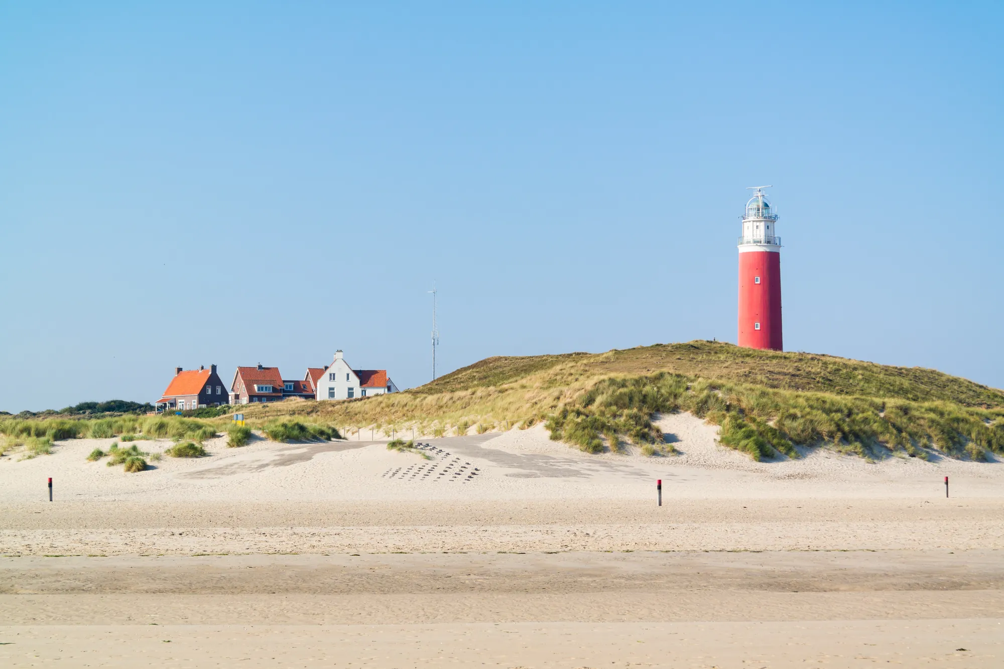 Roter Leuchtturm am Strand der Insel Texel, strahlt im klaren Himmel. Umgeben von feinem Sand und grünem Dünenbewuchs, bildet der Leuchtturm einen markanten Kontrast zum wolkenlosen Himmel.