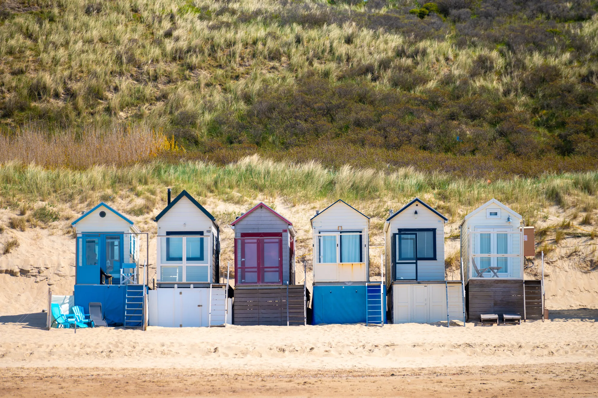 Das Bild zeigt eine Reihe bunter Badehäuser am Strand von Zoutelande auf Zeeland. Die kleinen, charmanten Häuschen in unterschiedlichen Farben stehen direkt am Sandstrand vor einer malerischen Dünenlandschaft. Jedes Badehaus hat eine eigene kleine Terrasse oder einen Sitzbereich, der die Nähe zum Meer betont. Die Dünen im Hintergrund sind mit Gras bewachsen, was der Szenerie eine natürliche, windgeschützte Atmosphäre verleiht. Der weiche Sand und die ruhige Umgebung strahlen eine entspannte, sommerliche Stimmung aus, typisch für die Küste von Zeeland.