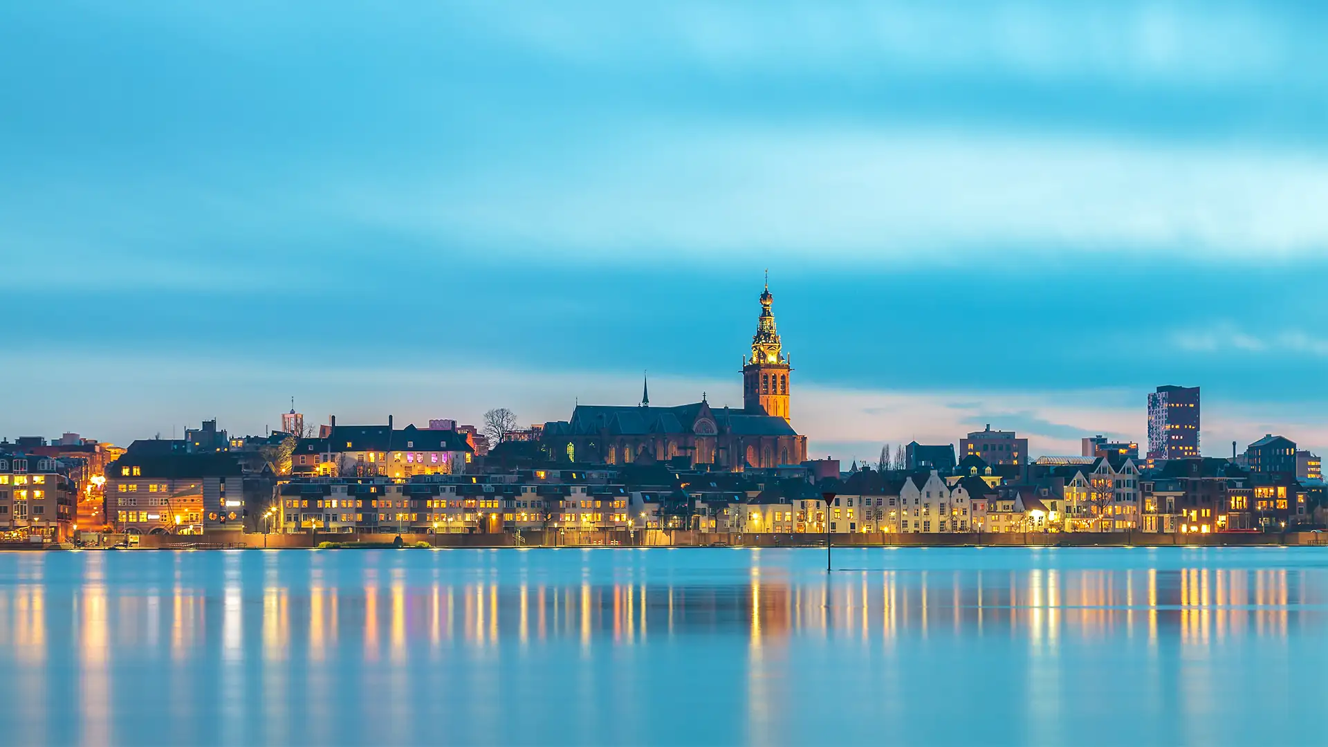 Panorama der Stadt Nijmegen mit beleuchteten Gebäuden und Kirchturm am Ufer des ruhigen Gewässers bei Abenddämmerung (im Kachelformat).