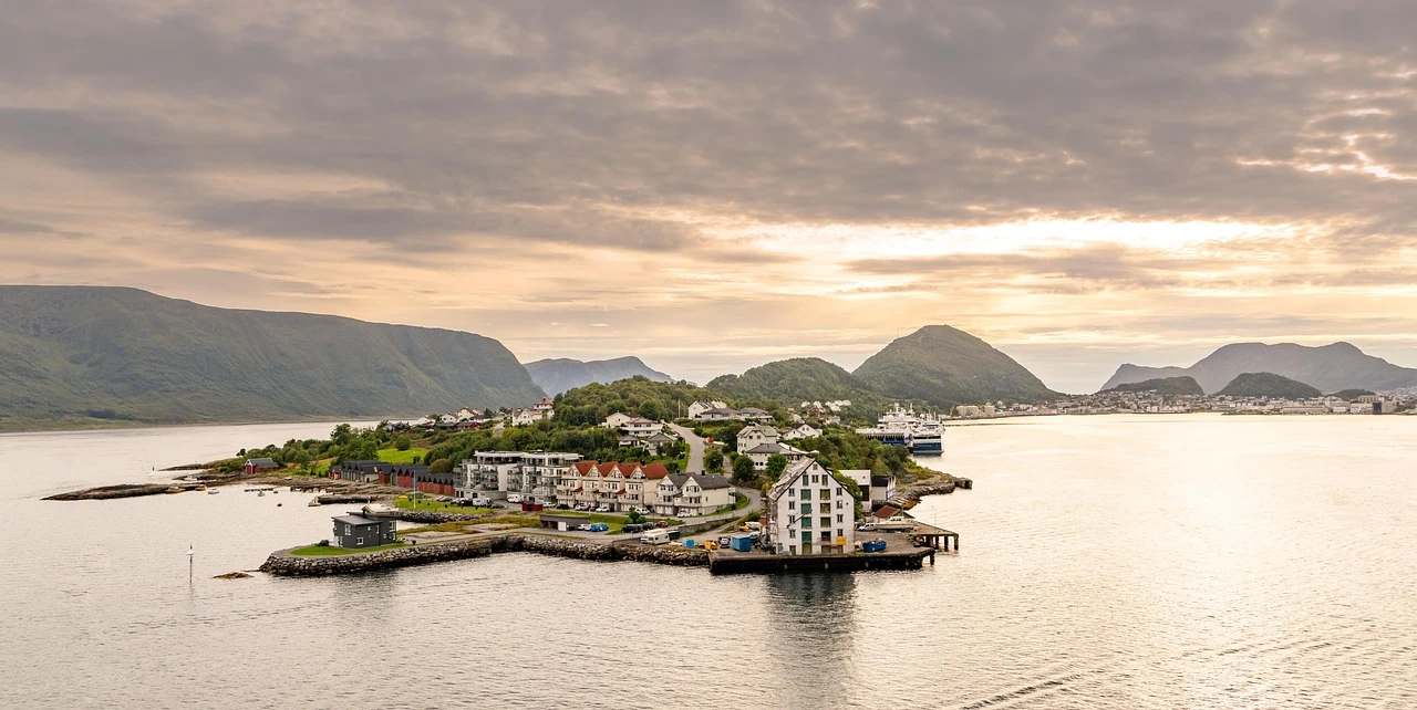 Ålesund_ Blick auf eine Insel mit mehreren Gebäuden, umgeben von Wasser und Bergen im Hintergrund, unter einem bewölkten Himmel.