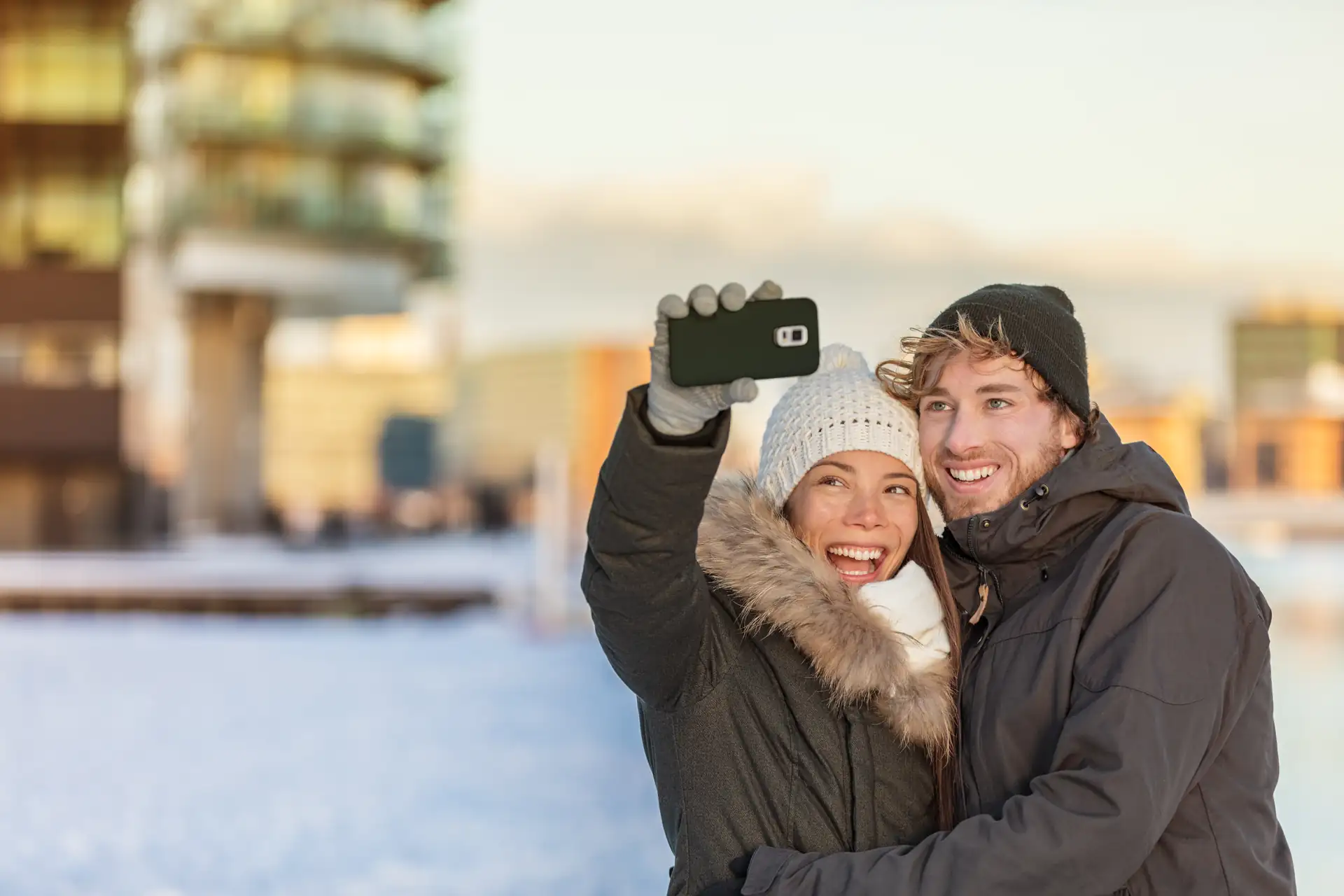 Zwei Personen in Winterkleidung machen ein Selfie im Freien, eine hält ein Smartphone mit schwarzer Hülle hoch.