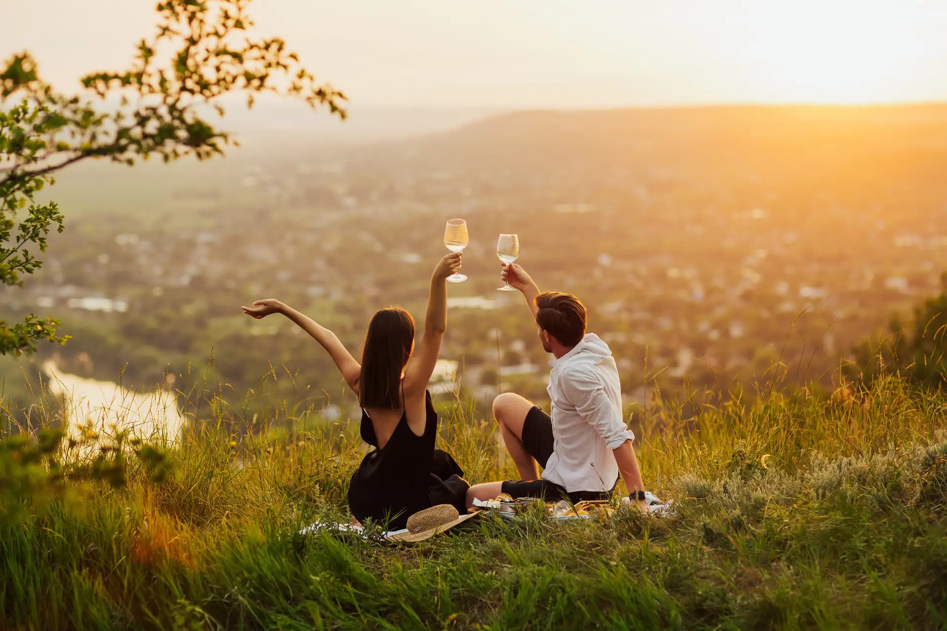 Paar sitzt auf einer Wiese mit Blick auf eine Stadt und stößt mit Weingläsern an