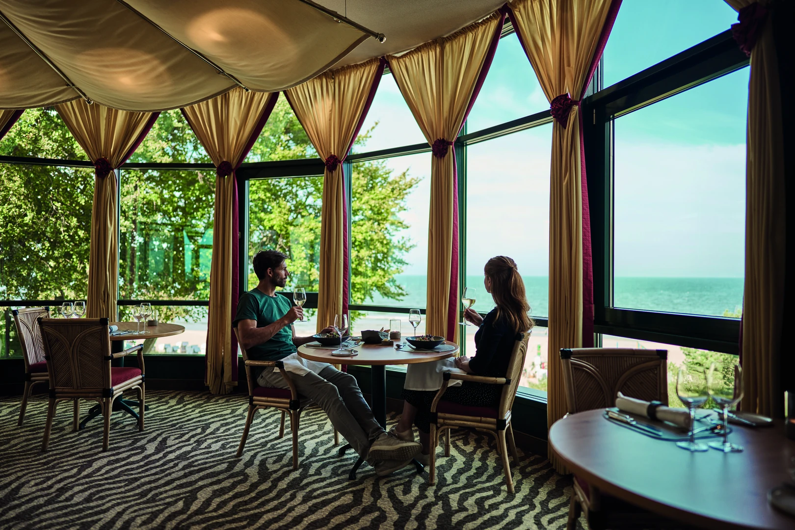 Ein eleganter Moment im Restaurant des aja Strandhotel Bansin: Gäste genießen ein stilvolles Dinner mit einem Glas Wein, während sie den Panoramablick auf die Ostsee durch die großen Fensterfronten bewundern. Die geschmackvolle Einrichtung und die entspannte Atmosphäre machen jede Mahlzeit zu einem besonderen Erlebnis.