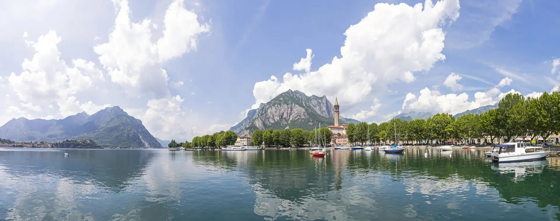 Panoramablick auf den Comer See bei Lecco mit Bergen, Uferpromenade, Bäumen und Booten auf dem Wasser