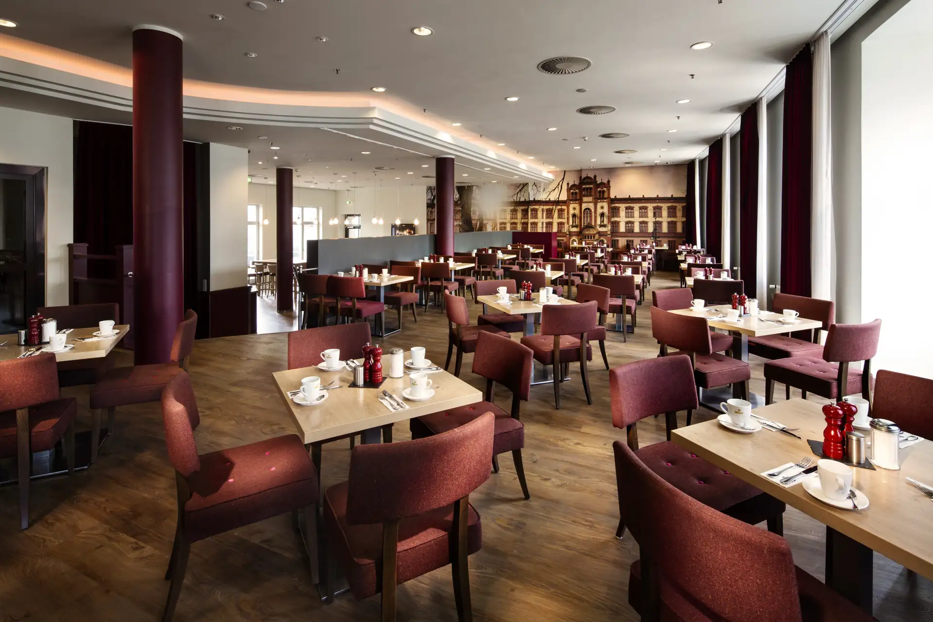 Großer, heller Speisesaal im Penta Hotel Rostock mit vielen Tischen und rotbraunen Stühlen, gedeckt mit Tassen und Tellern, und einem großen Wandbild im Hintergrund