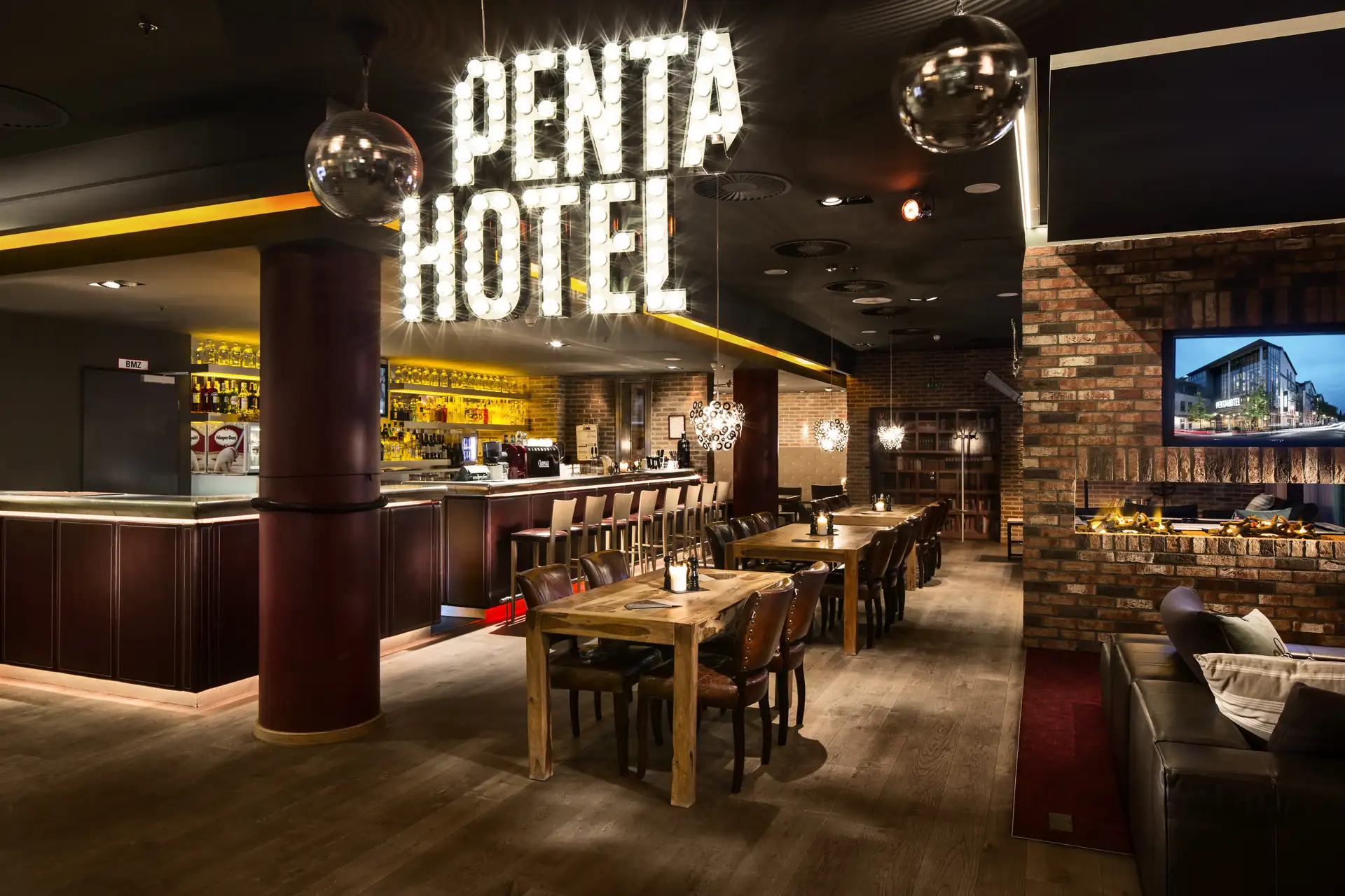 Lounge des Penta Hotel Rostock mit beleuchtetem Schriftzug 'PENTA HOTEL', Barbereich, Holztischen und Sitzgelegenheiten in warmem Ambiente