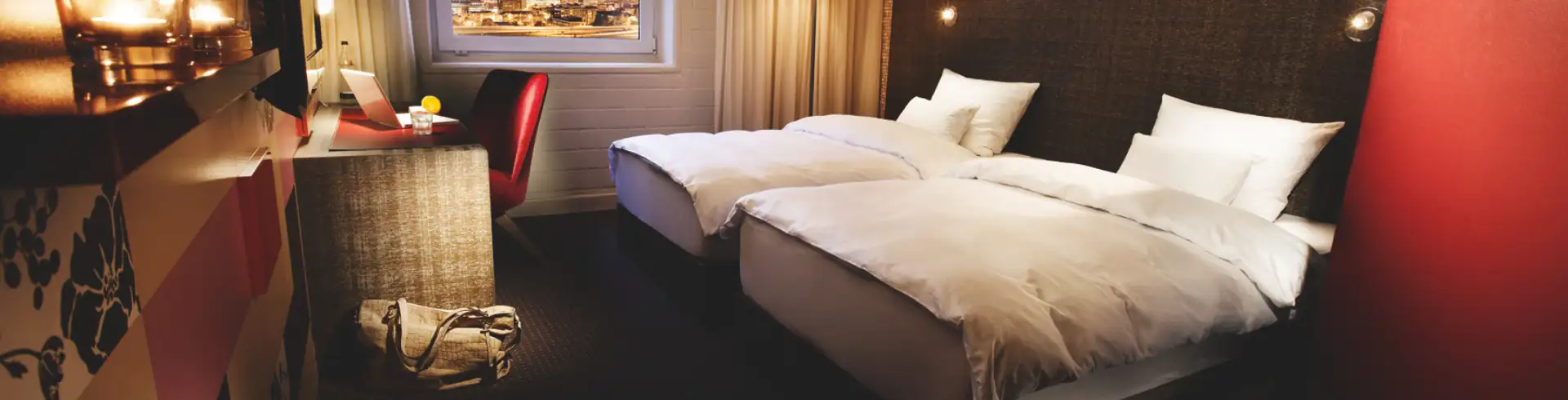 Hotelzimmer mit zwei Einzelbetten, Schreibtisch mit Stuhl vor Fenster, Stehlampe und dunklem Kopfteil mit indirekter Beleuchtung (im Bannerformat)