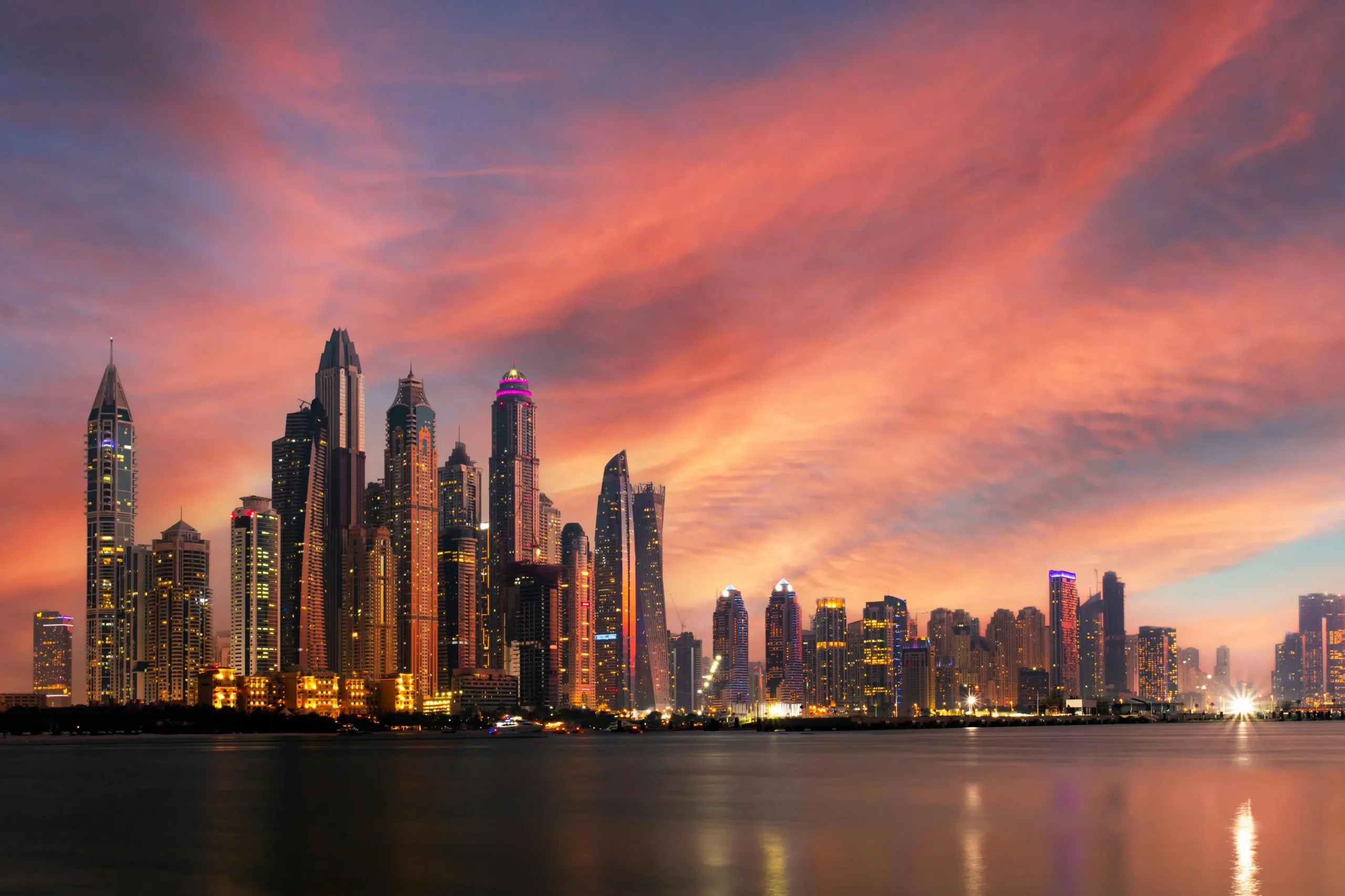 Dubai_Skyline einer modernen Stadt mit beleuchteten Wolkenkratzern bei Sonnenuntergang und orange-rotem Himmel über ruhigem Wasser