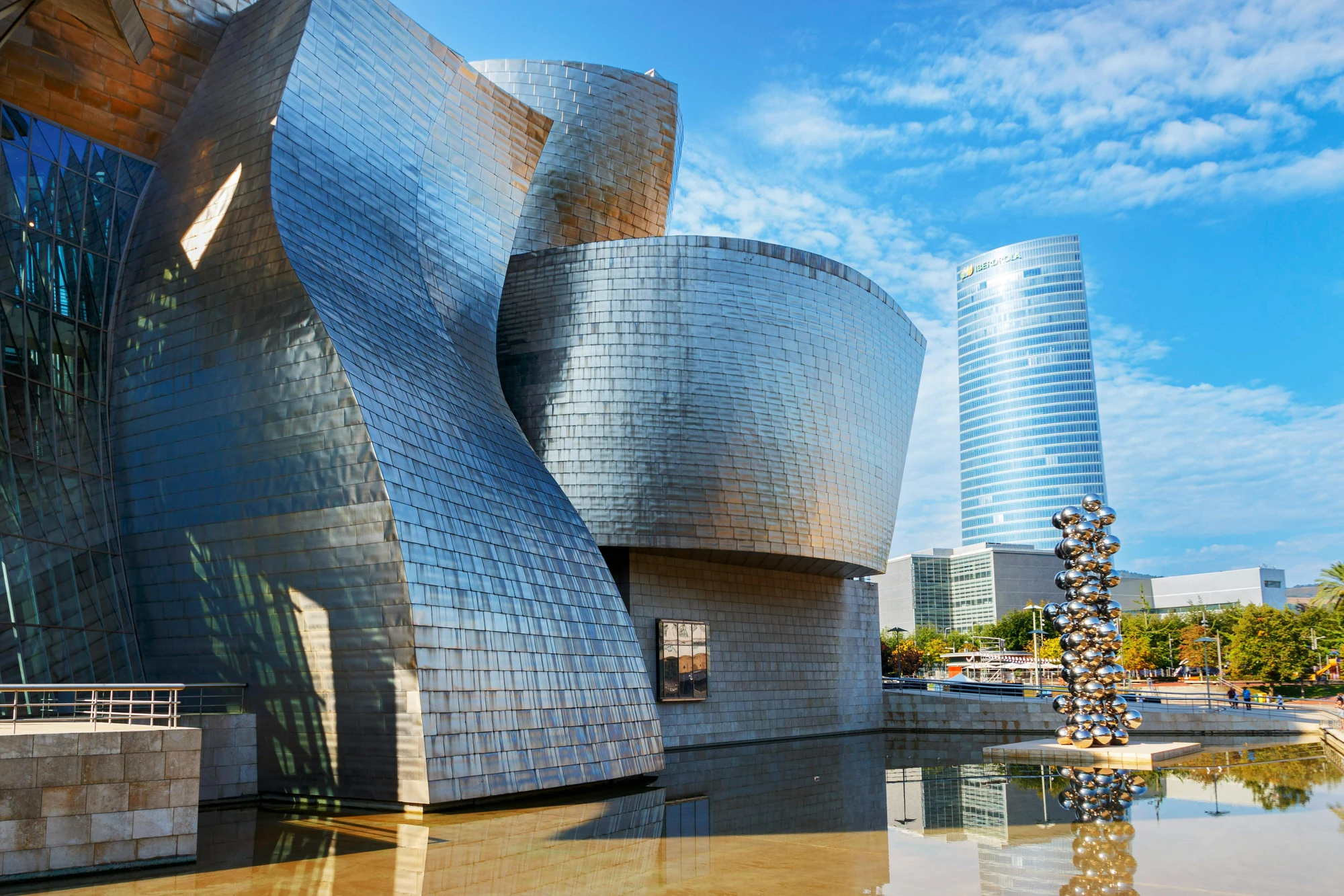 Das Guggenheim-Museum in Bilbao mit geschwungenen, metallischen Fassaden und einer reflektierenden Wasserfläche im Vordergrund. Im Hintergrund ein moderner Wolkenkratzer.