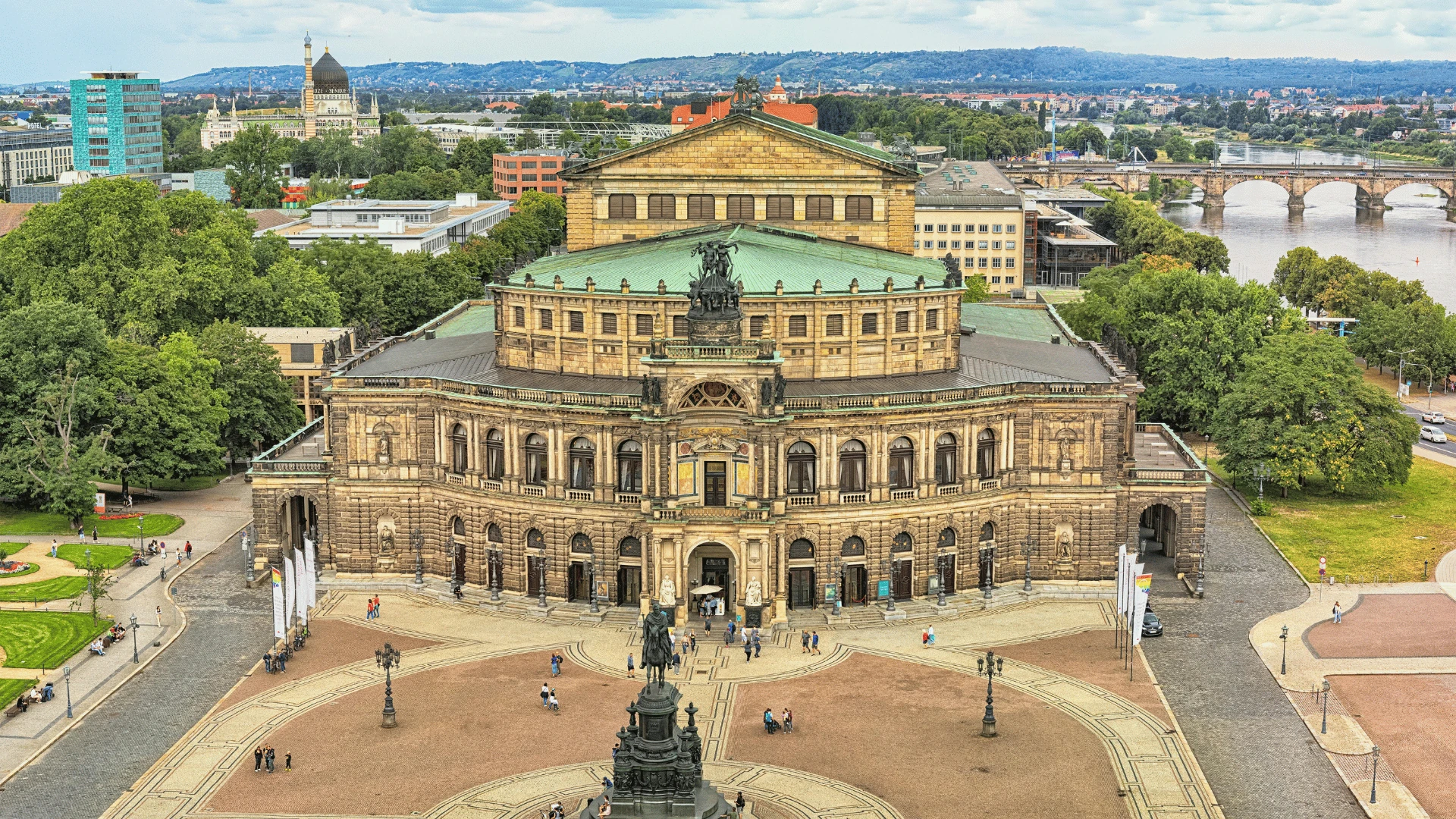 Die Semperoper in Dresden, eines der bekanntesten Opernhäuser der Welt, wird hier aus einer beeindruckenden Perspektive gezeigt. Der Blickfang dieser Aufnahme ist die prachtvolle Fassade des Gebäudes, das im Stil der Neorenaissance erbaut wurde. Im Vordergrund ist das Reiterstandbild von König Johann von Sachsen zu sehen, das vor dem Haupteingang der Oper aufgestellt ist. Der weite Platz vor der Semperoper lädt zu einem Spaziergang ein und bietet einen herrlichen Blick auf das Gebäude sowie auf die umliegende Architektur und die Elbe. In der Ferne ist die bekannte Augustusbrücke sichtbar, die über den Fluss führt. Die grüne Umgebung und die charakteristische Architektur der Stadt Dresden verleihen der Szene einen besonderen Charme.