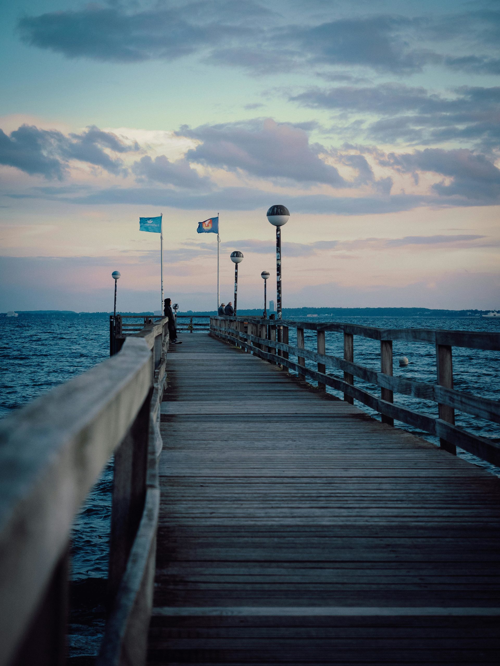 Ein hölzerner Steg ragt ins Wasser, Fahnen flattern und Menschen spazieren unter einem bunten, wolkenverhangenen Himmel in der Abenddämmerung.