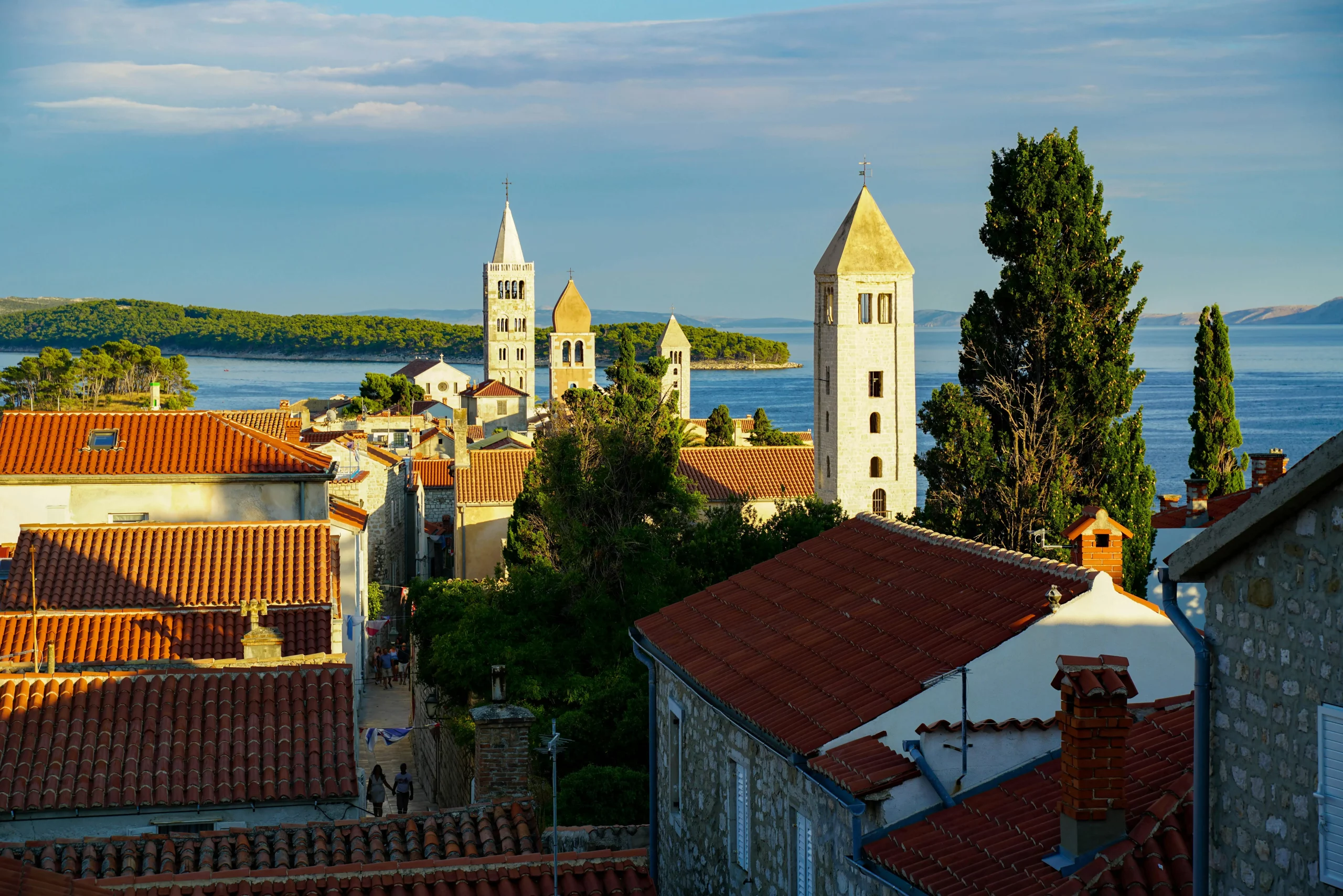 Blick auf die Altstadt von Rab mit mehreren Kirchtürmen und roten Ziegeldächern, dahinter die Adria und grüne Inseln