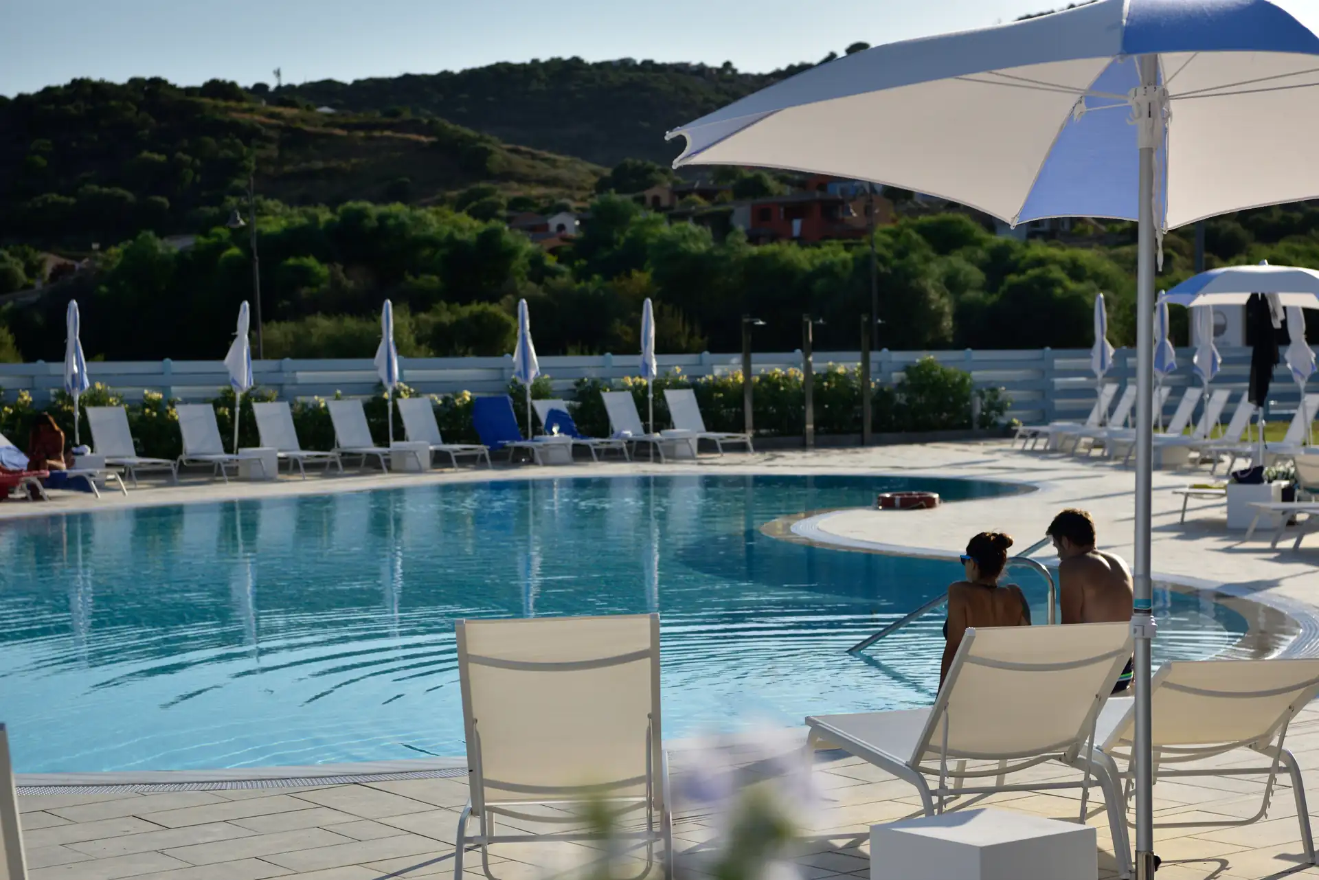 Zwei Personen sitzen auf Liegestühlen im Hotel Piccolo in Porto Ottiolu unter einem Sonnenschirm neben einem Pool mit weiteren Liegestühlen und grünen Hügeln im Hintergrund
