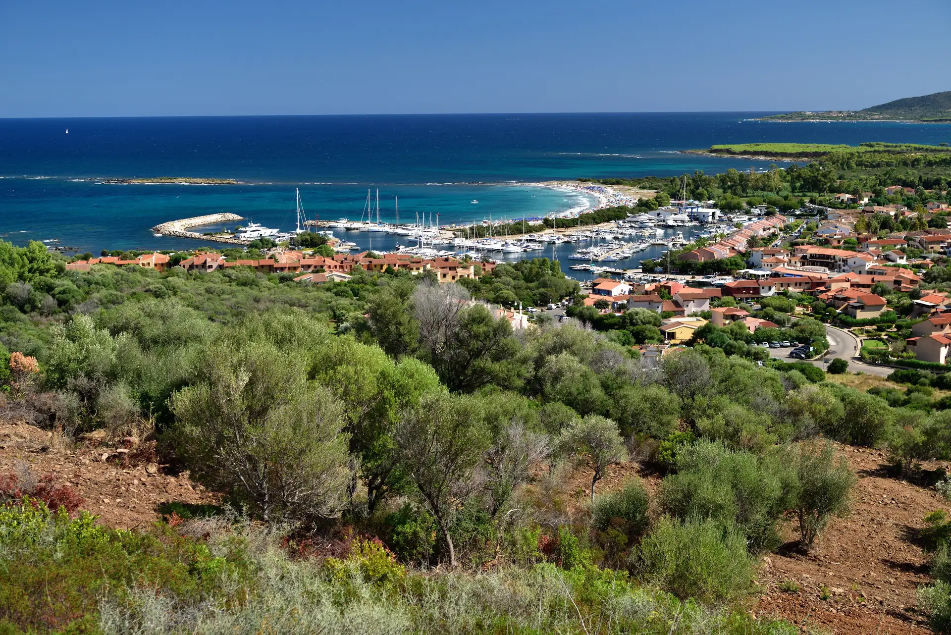 Blick auf Porto Ottiolu mit Hafen, Segelbooten und blauem Meer, umgeben von grünen Hügeln und Vegetation