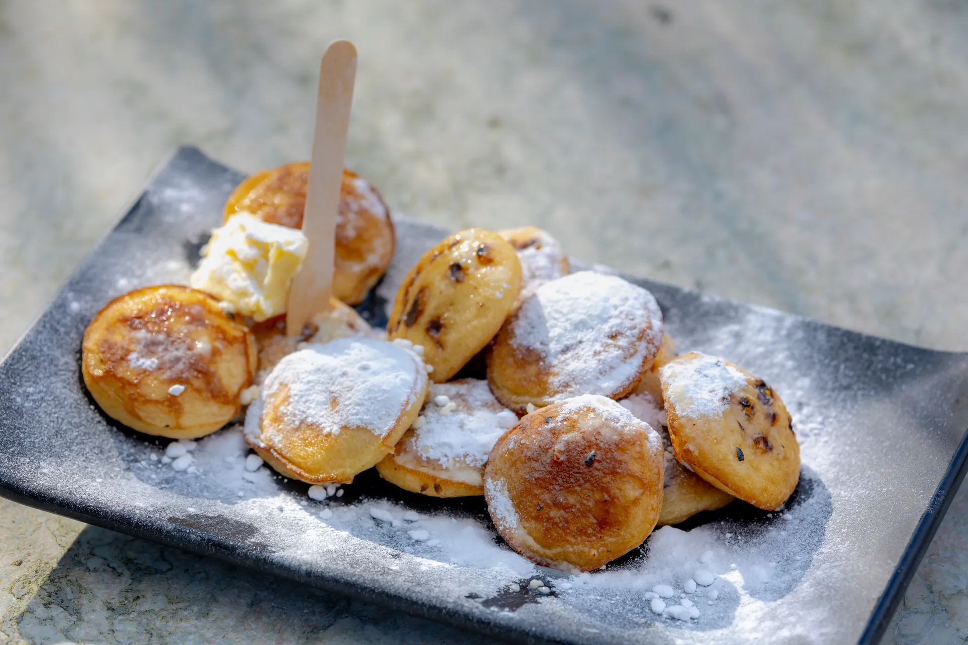 Nahaufnahme von frisch zubereiteten Poffertjes, kleinen, fluffigen niederländischen Pfannkuchen, bestäubt mit feinem Puderzucker. Die goldbraune Oberfläche und die zarten, runden Formen laden zum Genießen ein und verleihen dem Bild eine verlockende Ästhetik.