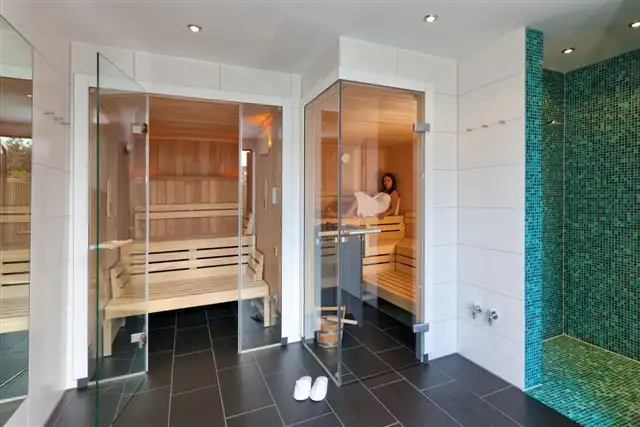 Saunabereich mit zwei Saunakabinen aus Holz und Glas, eine Kabine ist mit einer sitzenden Frau belegt, daneben ein kleiner Pool mit grünen Mosaikfliesen und weißen Hausschuhen auf dem Boden.