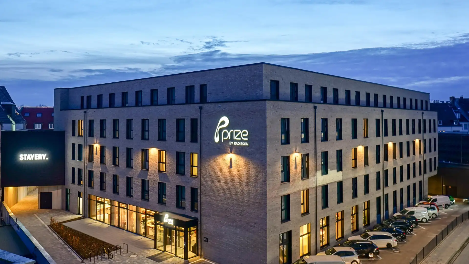 Prize by Radisson Osnabrück: Mehrstöckiges modernes Gebäude mit beleuchtetem Schriftzug 'Prize by Radisson' an der Fassade und mehreren geparkten Autos davor.