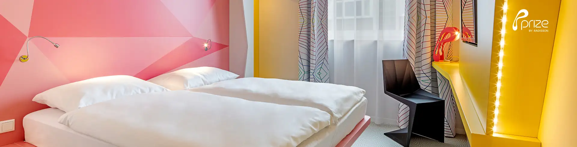 Prize Münster Banner: Modern eingerichtetes Hotelzimmer mit Doppelbett, gelben Wänden und schwarzem Stuhl vor Fenster