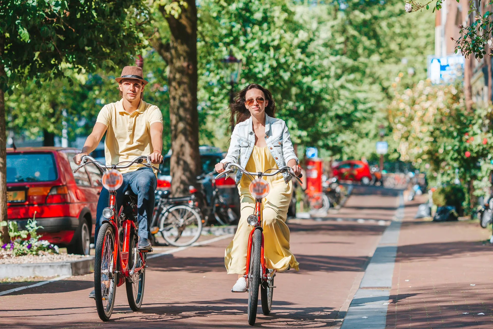 Pärchen radelt entspannt durch die malerischen Straßen Amsterdams an einem sonnigen Tag. Umgeben von historischen Gebäuden und grünen Bäumen genießen sie die frische Luft und die lebendige Atmosphäre der Stadt, während sie die Schönheit der Umgebung erkunden.
