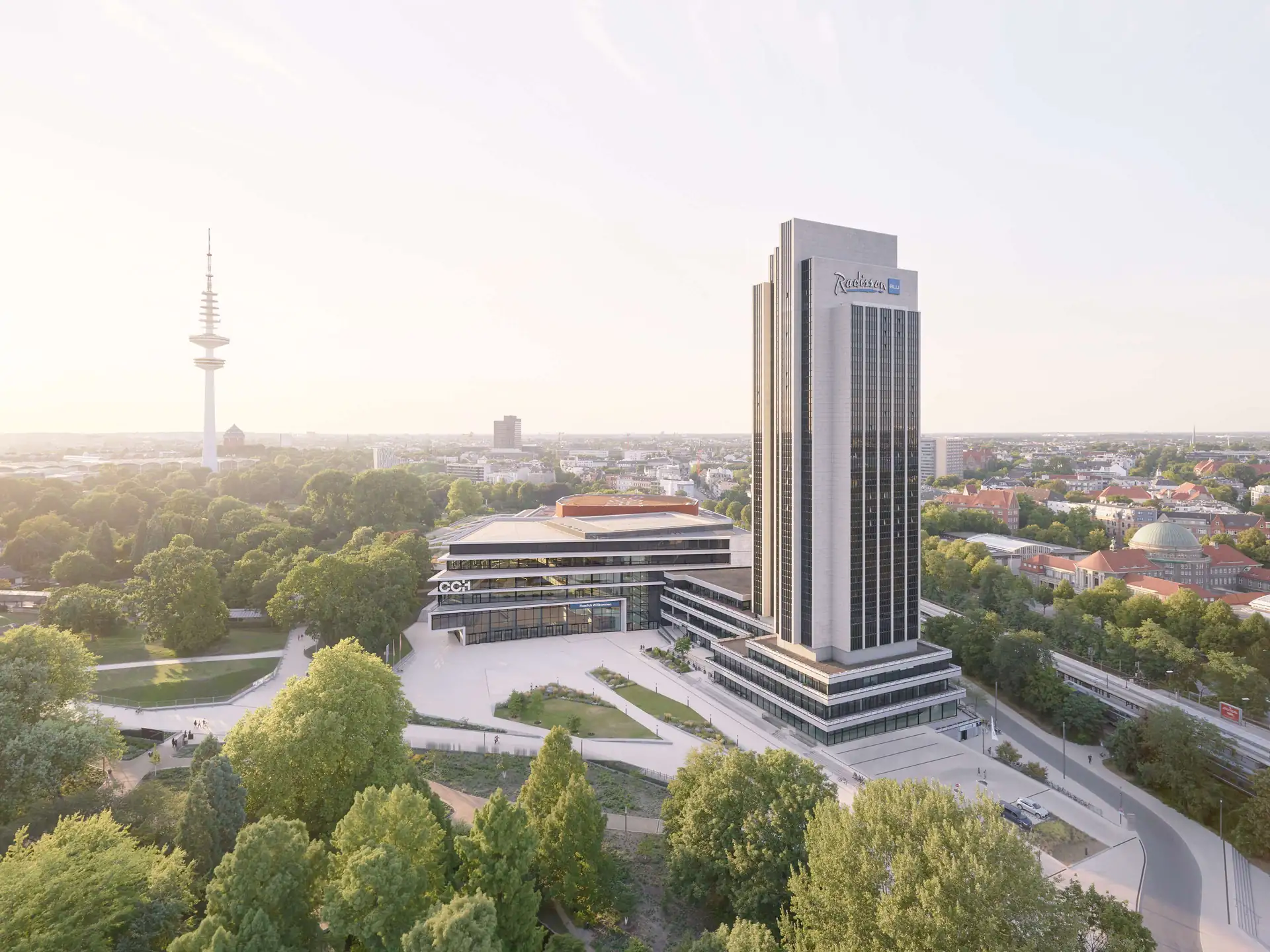 Hotel Radisson Blu Hamburg: Hochhaus mit dem Schriftzug 'Radisson Blu' neben einem flachen Gebäudekomplex umgeben von Bäumen und einer Stadtlandschaft im Hintergrund