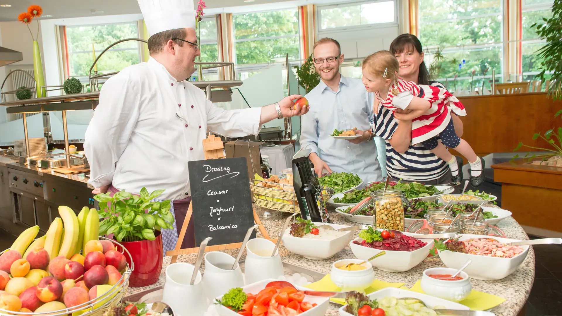 Koch in weißer Uniform reicht Gästen mit Kind frisches Gemüse am Buffet mit Salaten und Obst in einem hellen Speiseraum im Seminaris SeeHotel Potsdam
