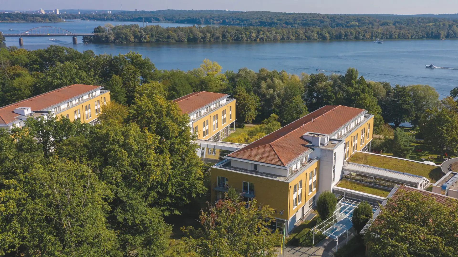 Gelbe Gebäude mit roten Dächern vom Seminaris SeeHotel Potsdam umgeben von Bäumen am Ufer des Templiner Sees, im Hintergrund Brücke und Wald