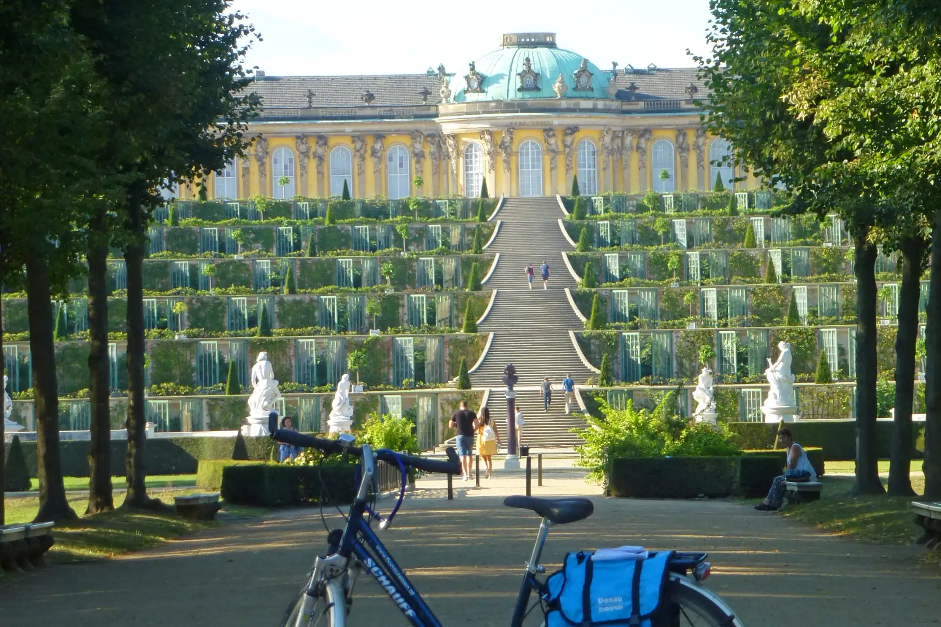 Radreise Potsdam (Bild im Kachelformat): Fahrrad mit blauer Tasche im Vordergrund, im Hintergrund Schloss Sanssouci mit Terrassengarten und Treppe, umgeben von Bäumen