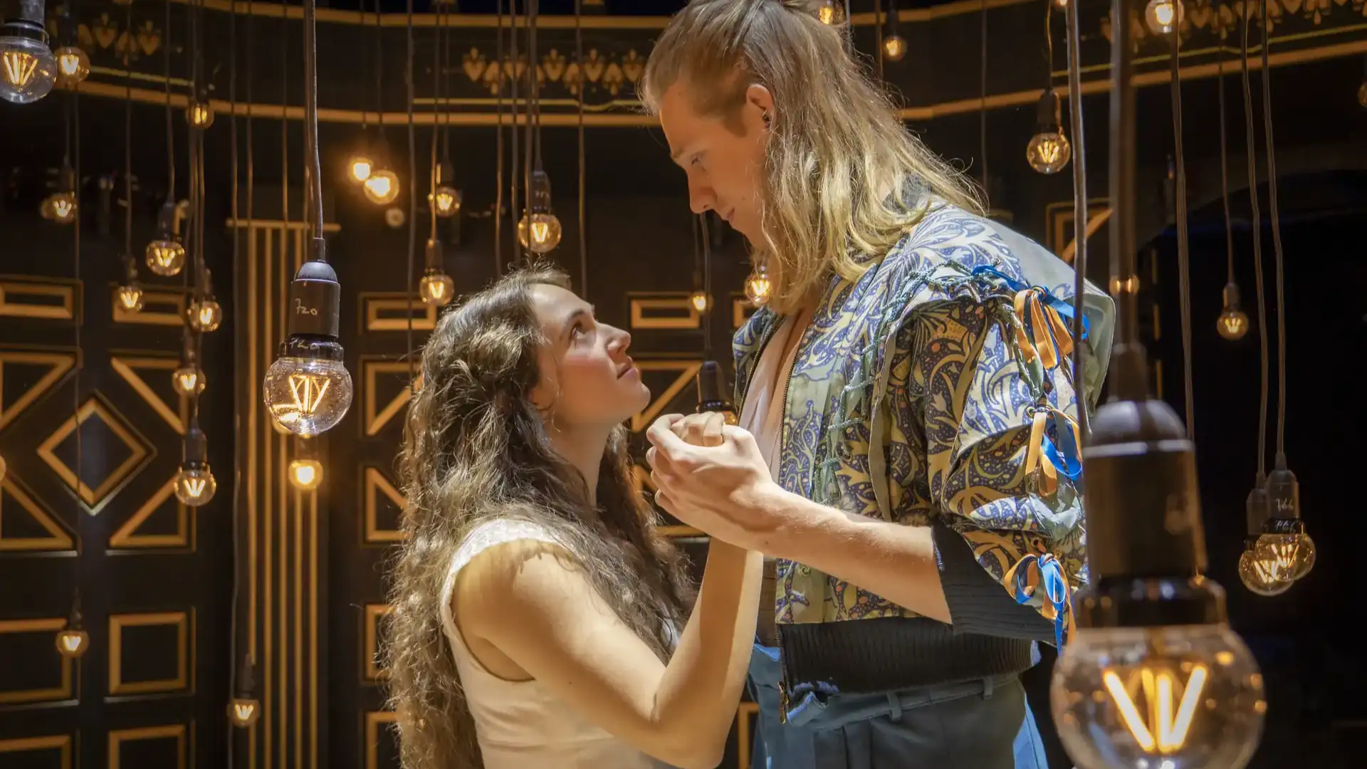 Romeo & Julia – Das Musical: Zwei Schauspieler in historischen Kostümen halten sich an den Händen, umgeben von hängenden Glühbirnen in einem dunklen Raum mit goldenen Verzierungen.