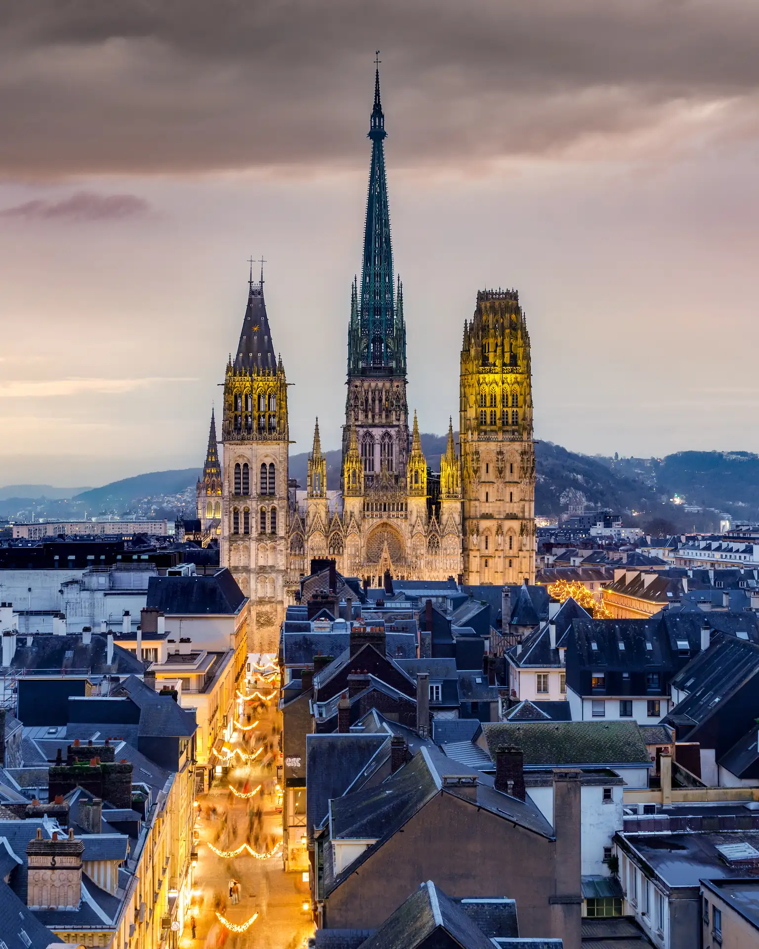 Rouen: Gotische Kathedrale mit drei beleuchteten Türmen in einer Stadt bei Dämmerung, umgeben von Häusern und Straßen.