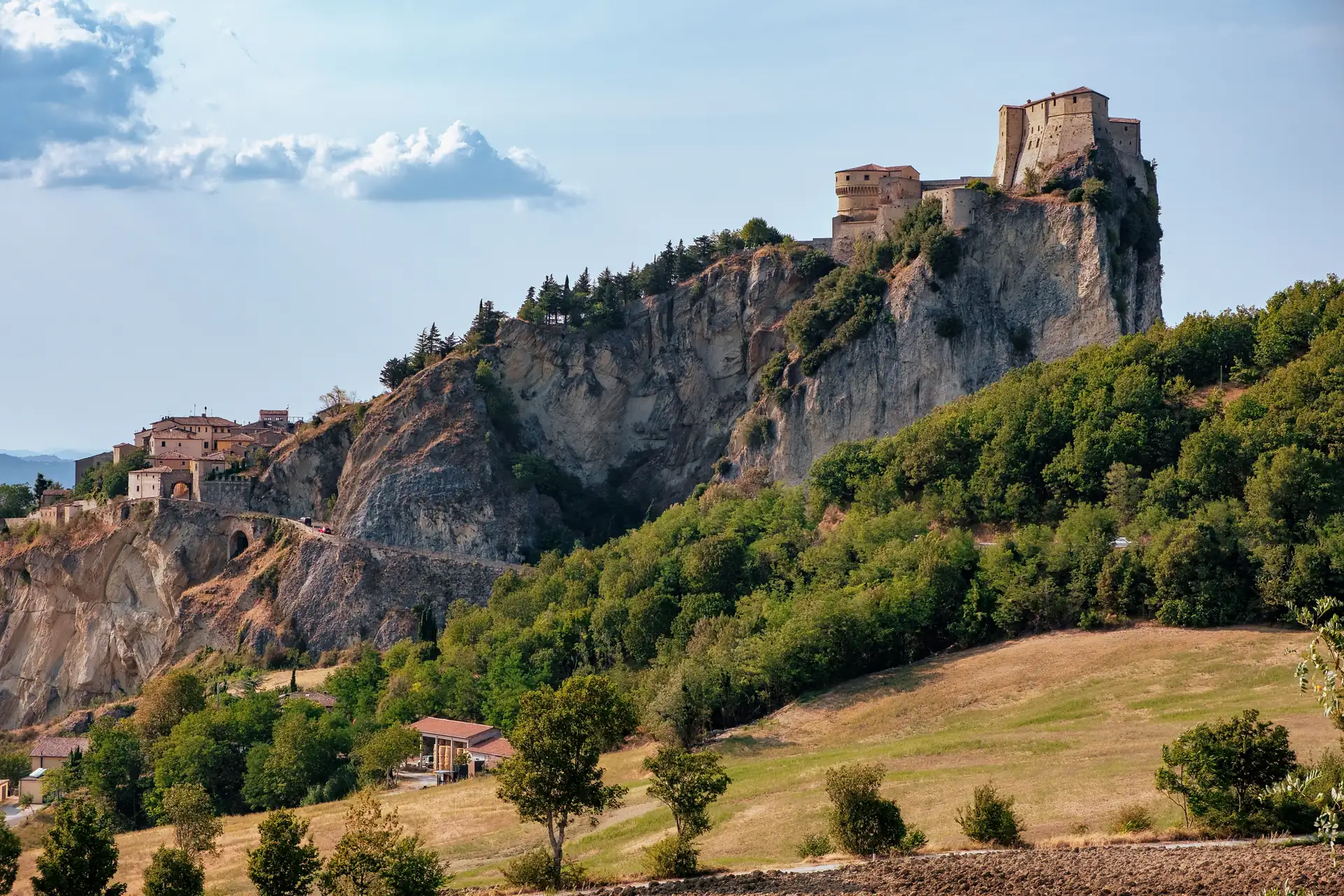 San Leo mit Burg auf einem steilen Felsen über bewaldeten Hügeln und Feldern unter blauem Himmel mit Wolken in der Emilia Romagna.
