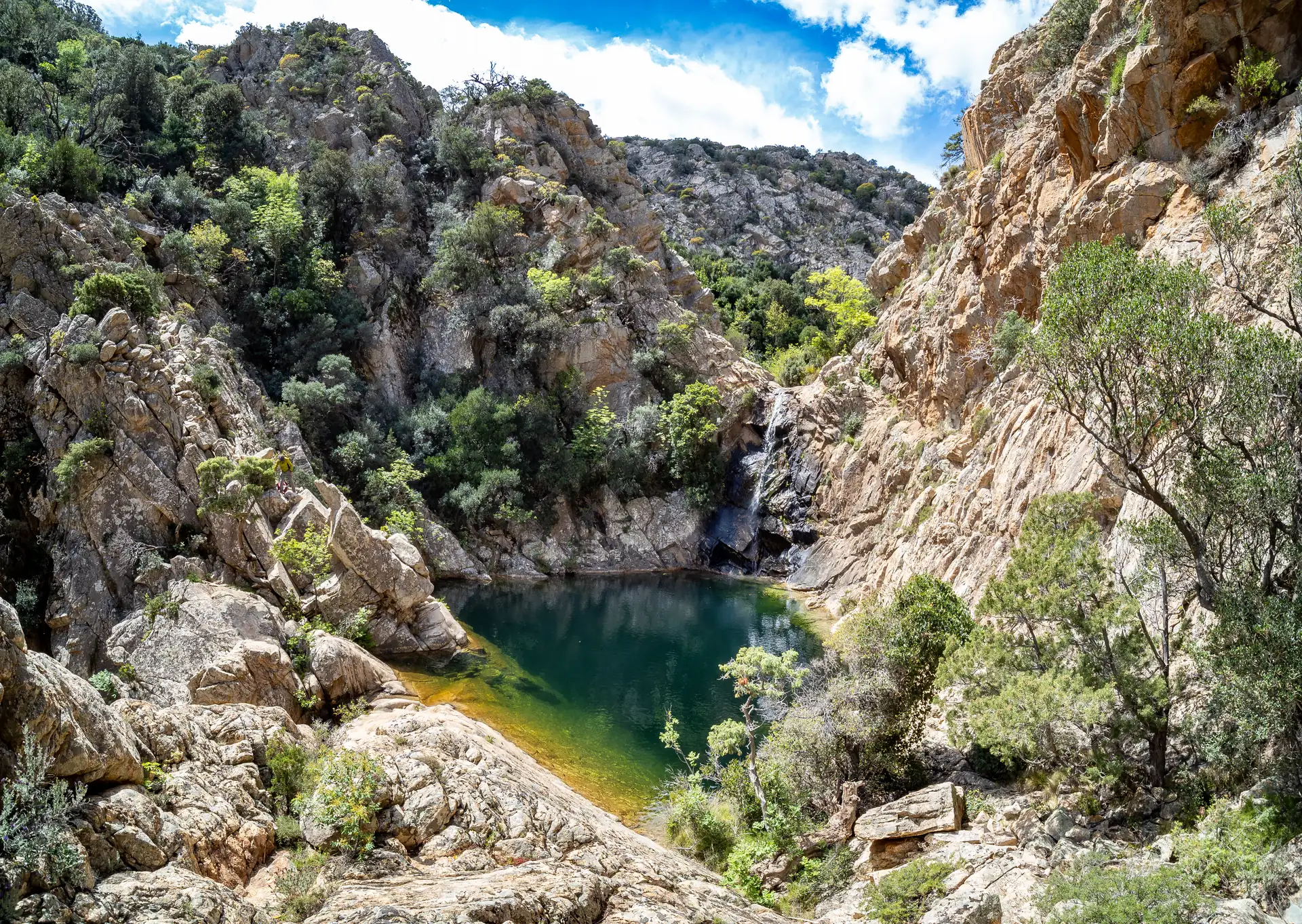 Grüner Bergsee umgeben von felsigen Klippen und bewaldeten Hängen unter blauem Himmel mit Wolken am Monte Nieddu auf Sardinien
