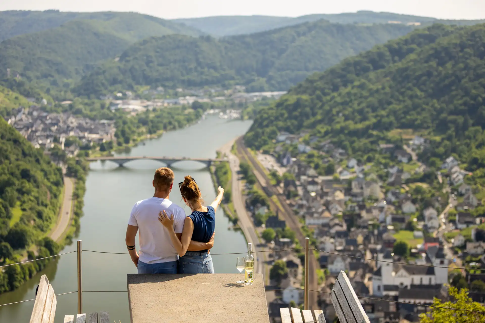 Paar sitzt auf einer Aussichtsplattform mit Blick auf die Mosel, umgeben von bewaldeten Hügeln und einer Brücke über den Fluss
