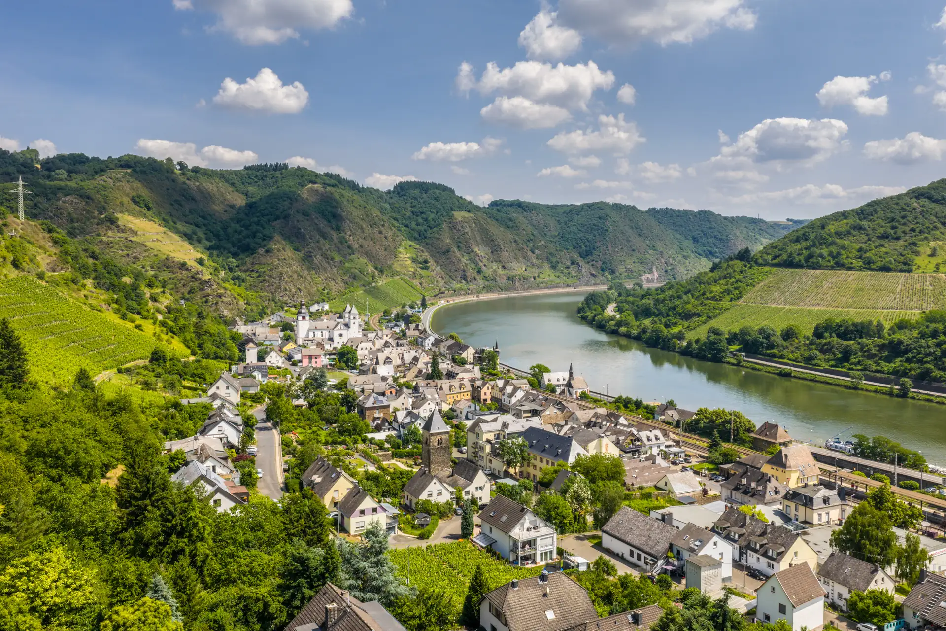 Blick auf Treis-Karden an der Mosel, umgeben von bewaldeten und bewirtschafteten Hügeln unter blauem Himmel mit Wolken