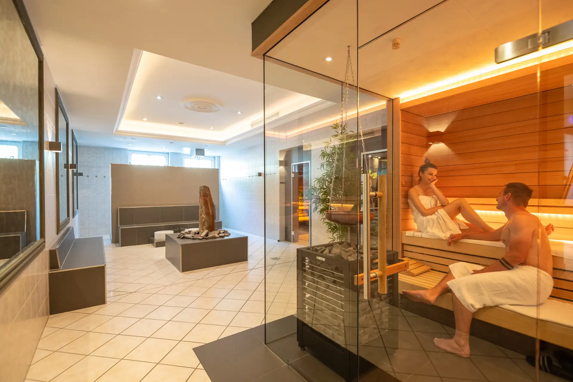 Wellnessbereich im Schloß-Hotel Petry mit Sauna aus Holz, in der ein Mann und eine Frau in weißen Handtüchern sitzen, davor ein moderner Ruheraum mit Stein-Dekoration und großen Fenstern