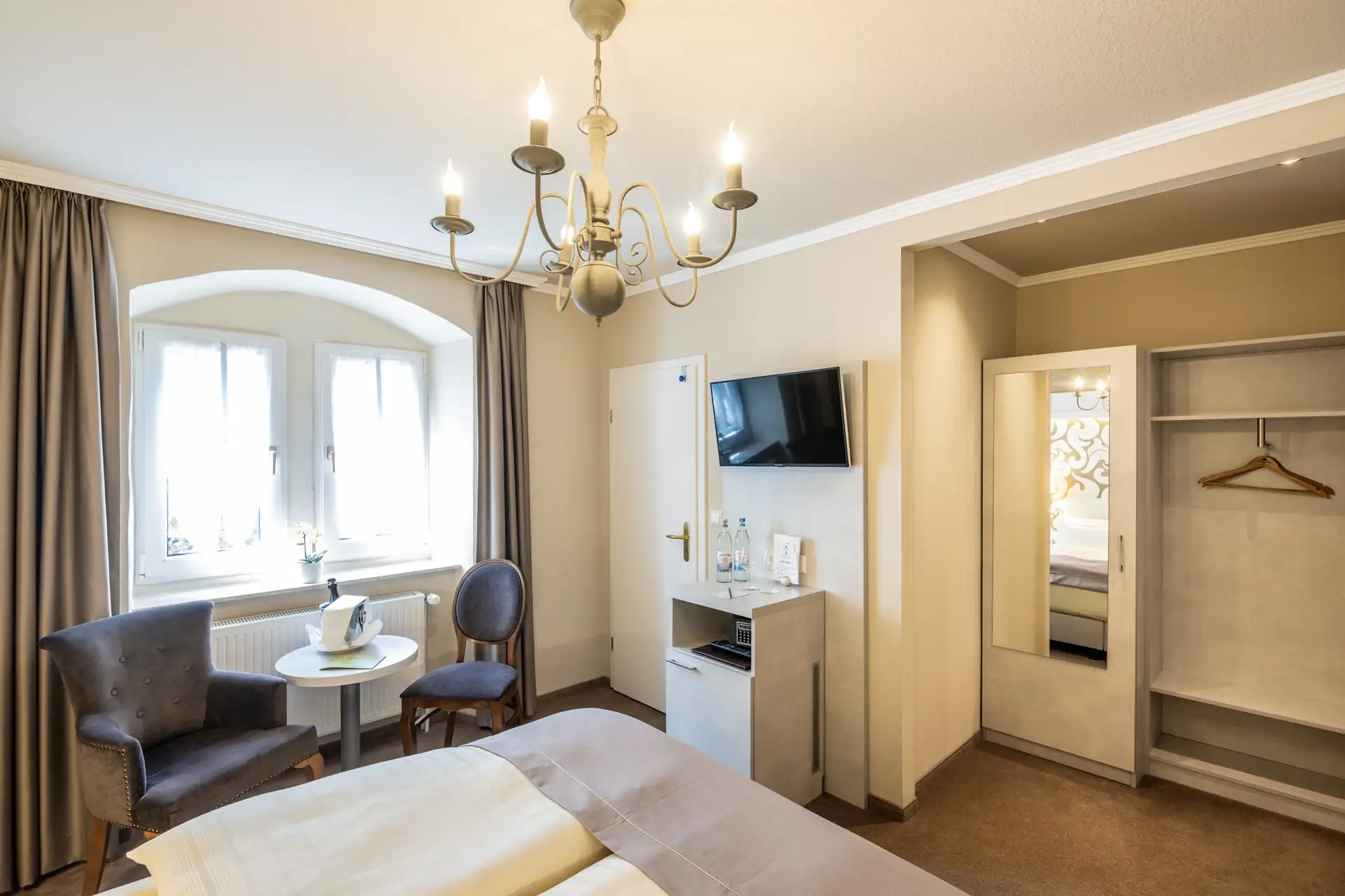 Hotelzimmer mit Doppelbett, zwei Sesseln und kleinem runden Tisch vor einem Fenster, Wand mit Flachbildfernseher und offenem Kleiderschrank mit Spiegel im Schloß-Hotel Petry