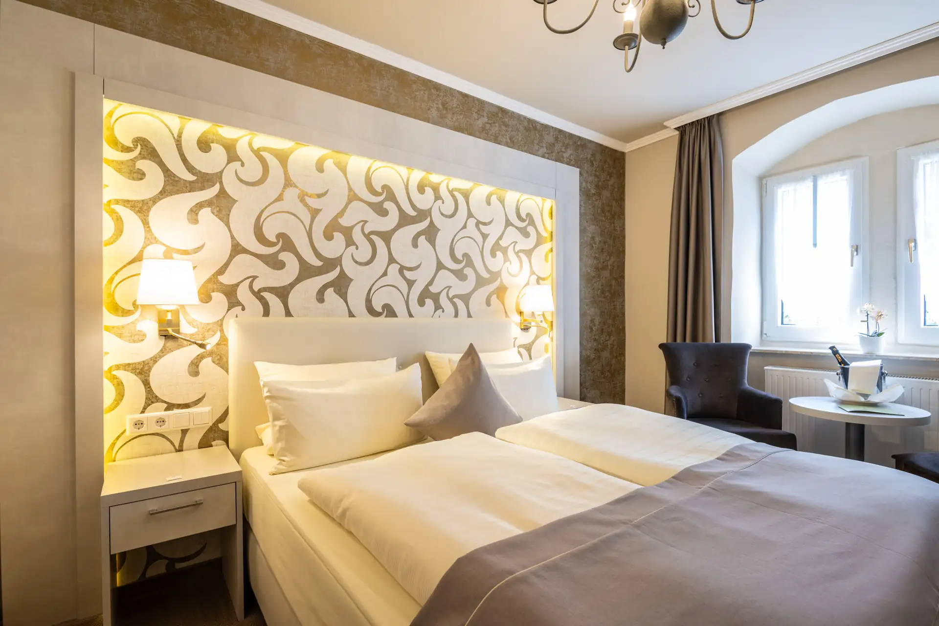 Hotelzimmer mit Doppelbett, dekorativem beleuchtetem Kopfteil, Nachttisch mit Lampe, Sessel und kleinem Tisch vor einem Fenster mit Vorhängen