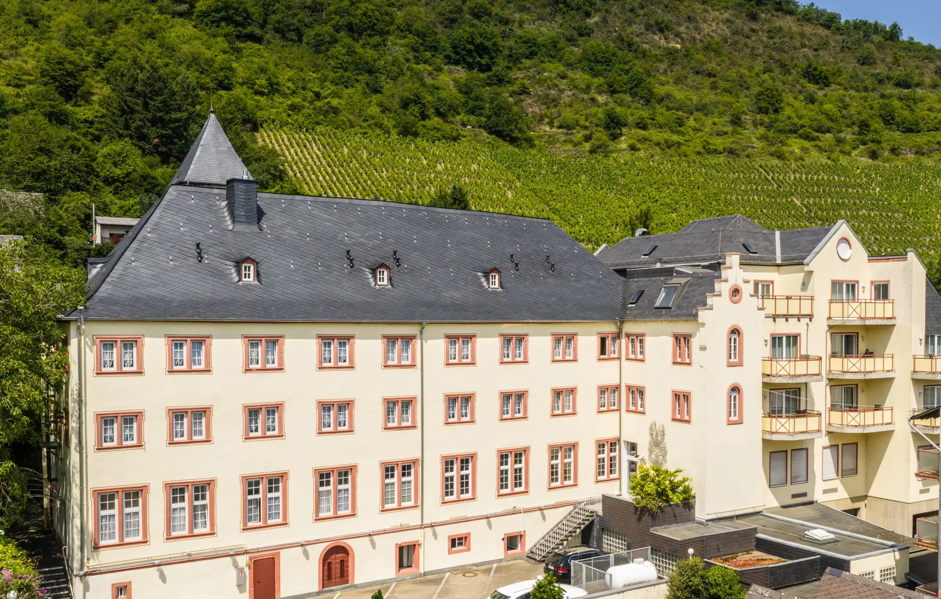 Das Schloß-Hotel Petry zeigt ein großes, helles Gebäude mit vielen Fenstern, einem spitzen Turm und einem bewaldeten Hügel im Hintergrund