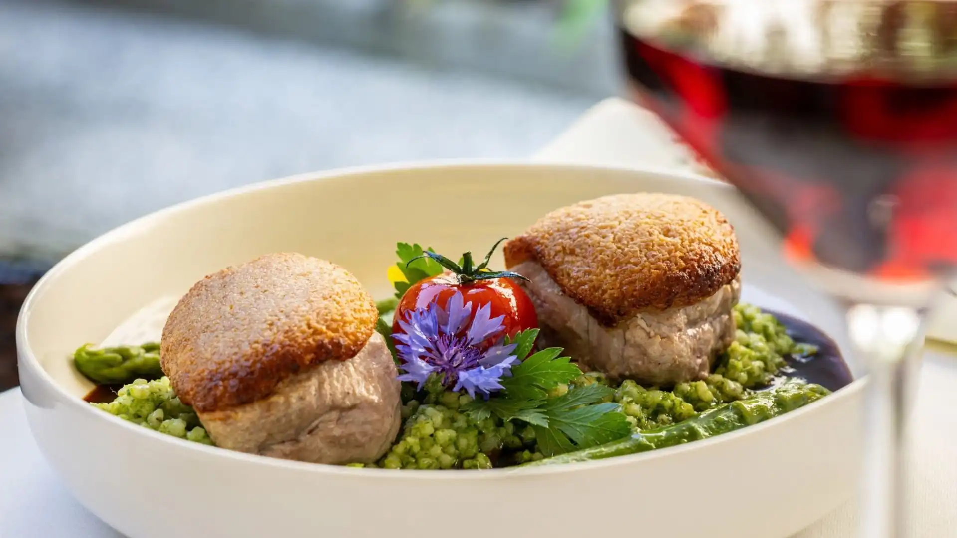 Zwei gebratene Fleischstücke auf grünem Gemüse mit einer Tomate und essbaren Blüten in einer weißen Schale