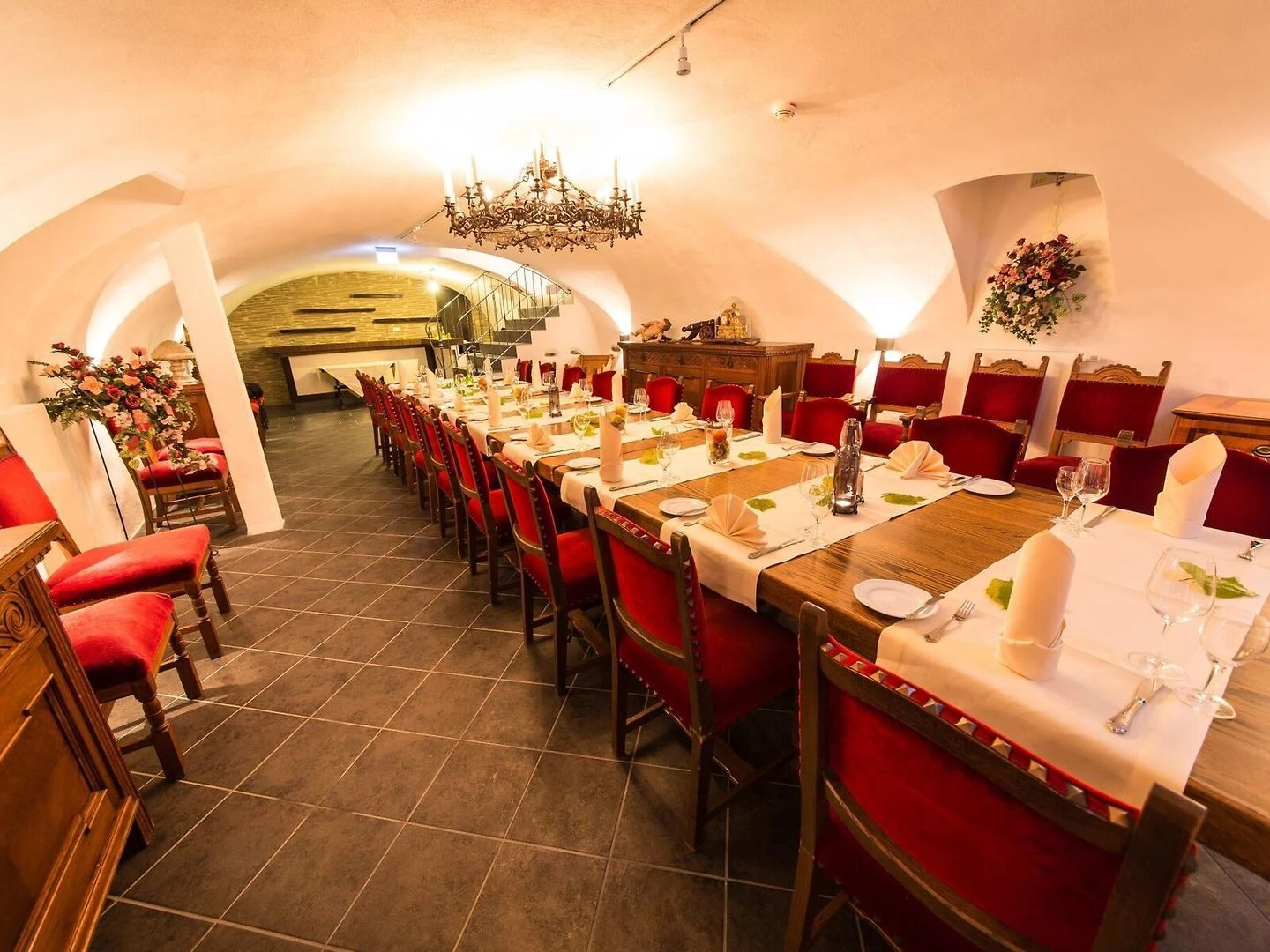 Langer gedeckter Esstisch mit roten Stühlen in einem Gewölbekeller mit Kronleuchter und Blumenarrangements