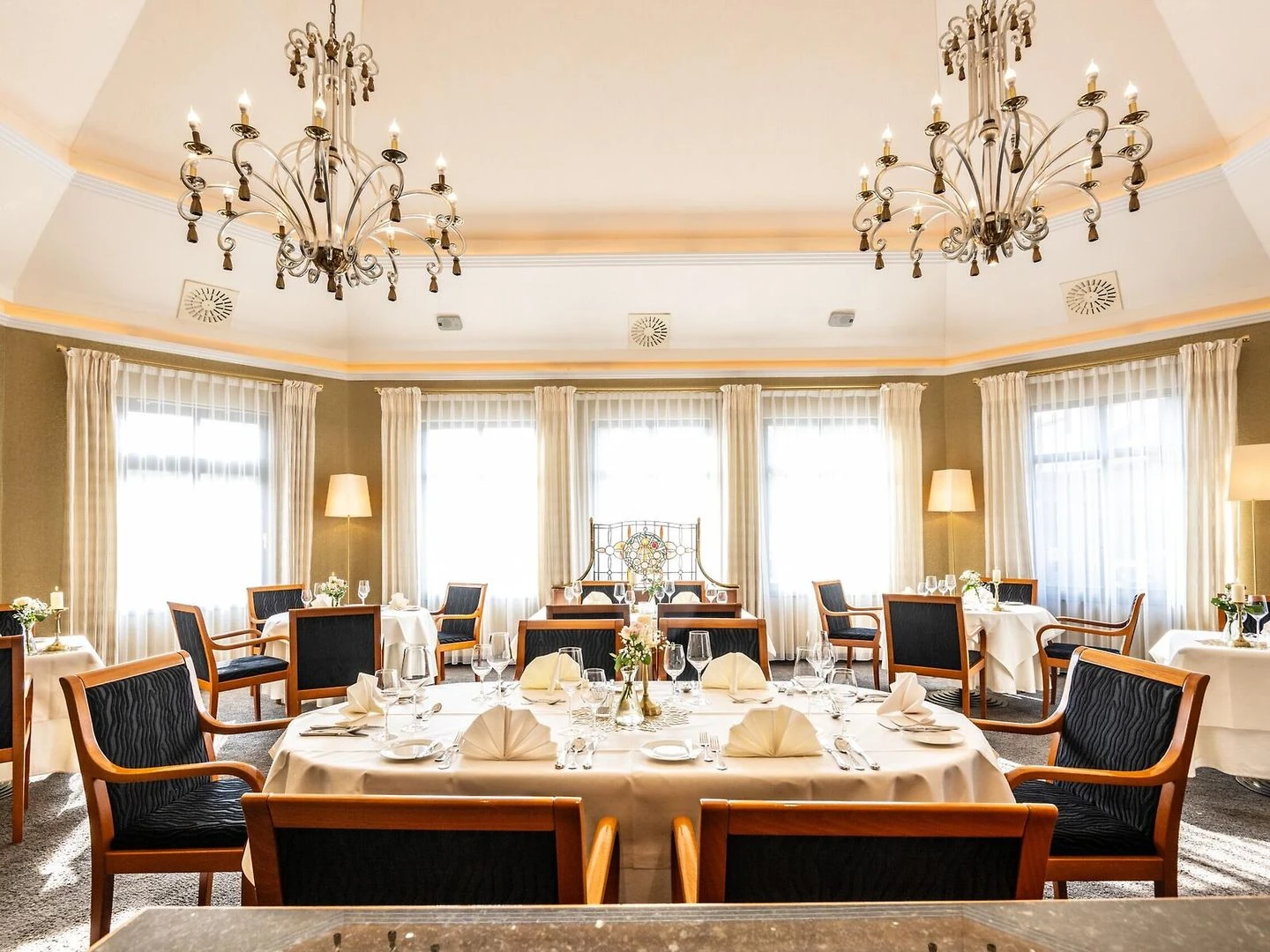 Eleganter Speisesaal mit runden Tischen, gedeckten weißen Tischdecken, Holzstühlen mit dunklen Polstern, großen Fenstern mit hellen Vorhängen und zwei Kronleuchtern an der Decke
