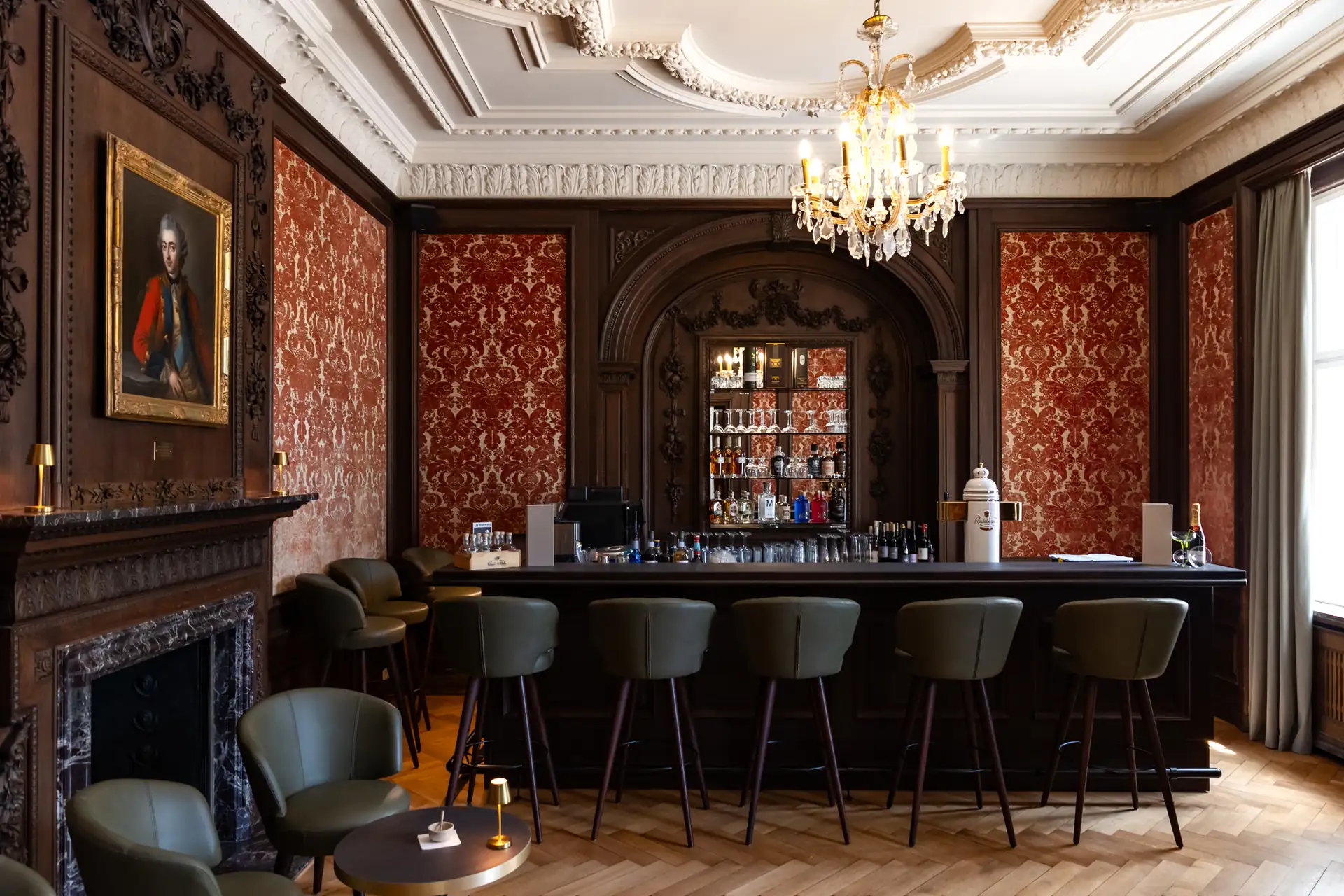 Elegante Hotelbar mit dunklem Holz, sechs Barhockern, Kronleuchter und rotem Tapetenmuster im Schlosshotel Neustrelitz