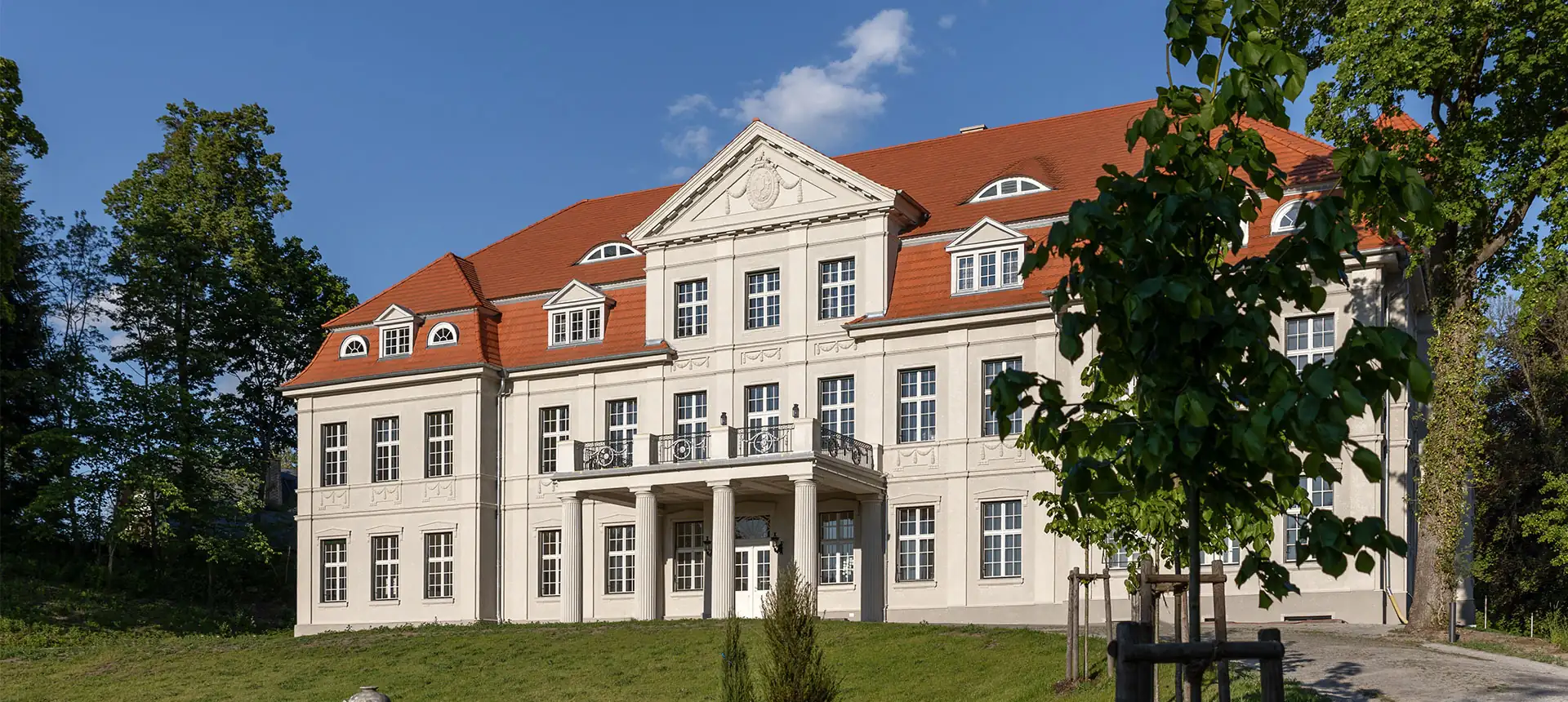 Schlosshotel Neustrelitz mit klassizistischer Fassade, dreigeschossig, rotem Satteldach und Säulenportikus, umgeben von Rasen und Bäumen (1920x860px)