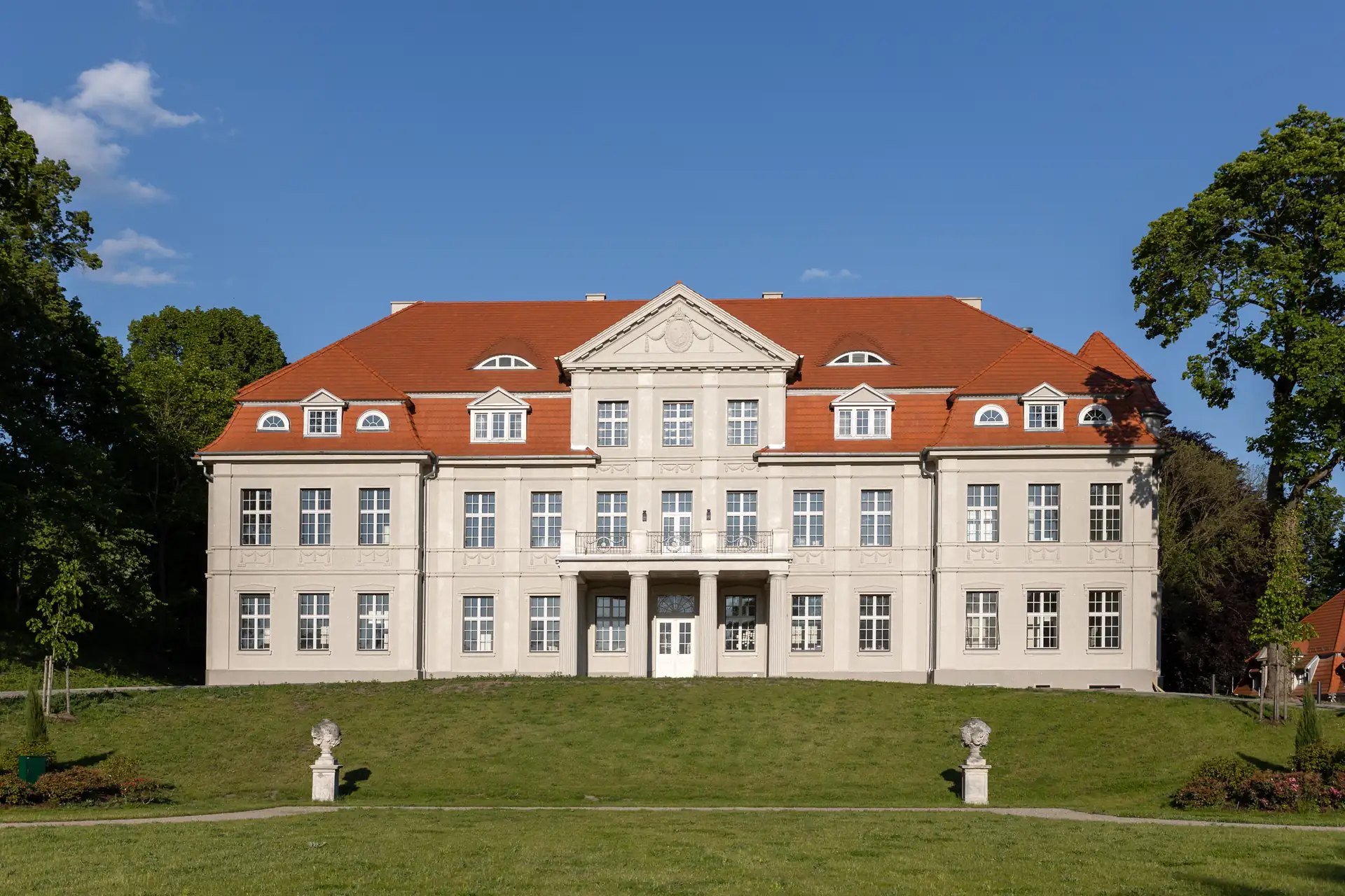 Frontalansicht des Schlosshotels Neustrelitz mit rotem Satteldach, symmetrischer Fassade und zwei steinernen Skulpturen auf dem Rasen davor