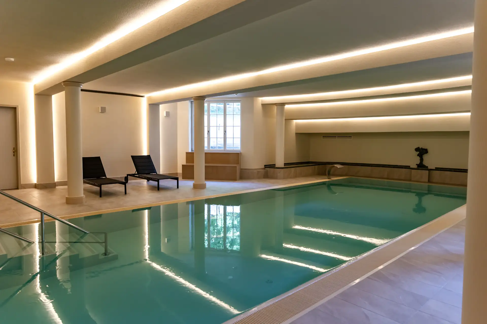 Innenpool des Schlosshotels Neustrelitz mit rechteckigem Becken, zwei schwarzen Liegen und großen Fenstern in einem hellen Raum mit Säulen