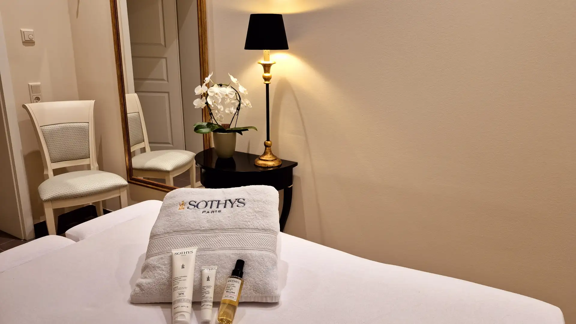 Weißes Handtuch mit schwarzem SOTHYS-Logo und drei Kosmetikprodukten auf einem weißen Bett in einem elegant eingerichteten Raum mit Stuhl, Spiegel, Nachttisch, Lampe und Orchidee