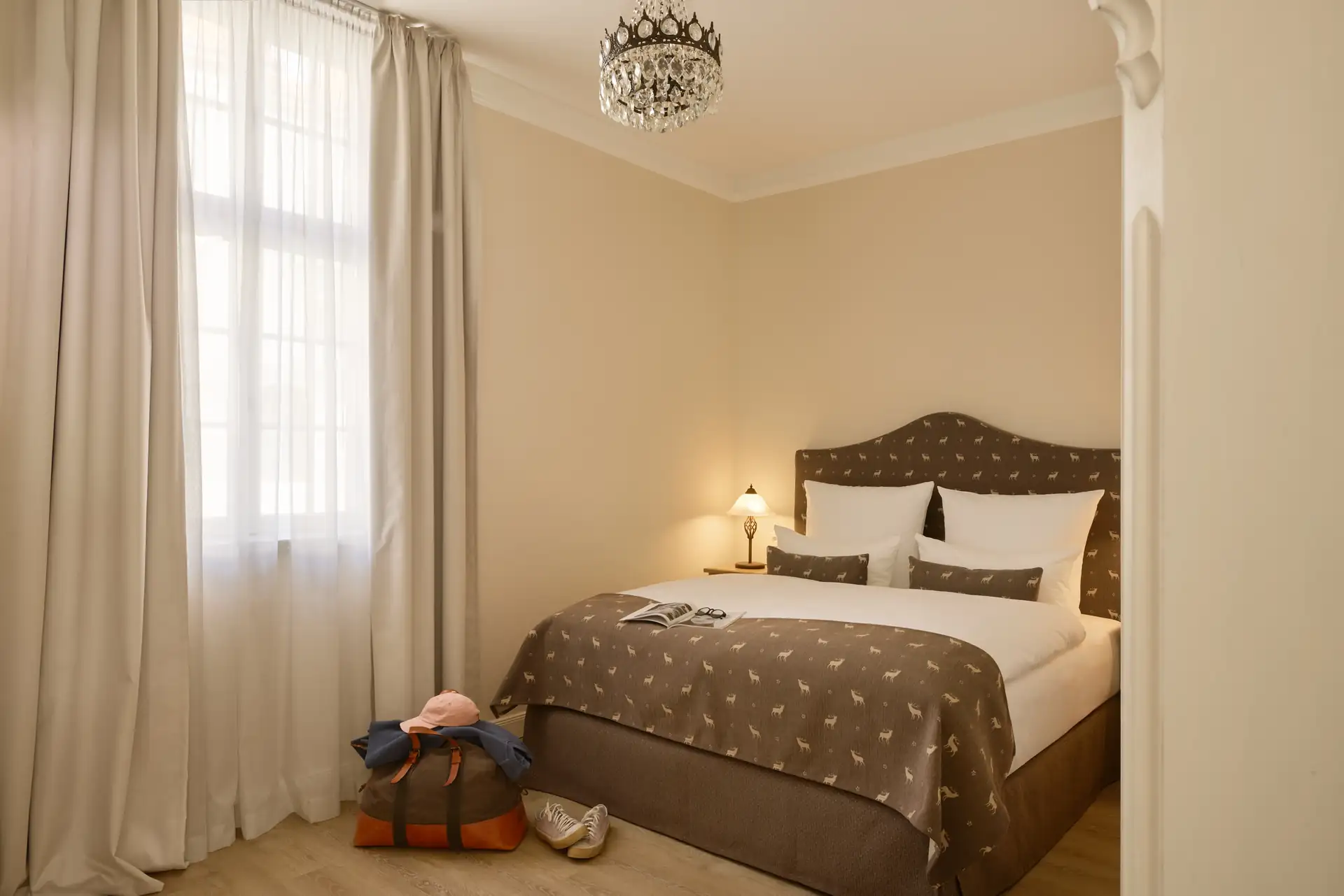 Hotelzimmer mit Doppelbett, gepolstertem Kopfteil, Nachttischlampe, Kronleuchter und Reisetasche auf dem Boden