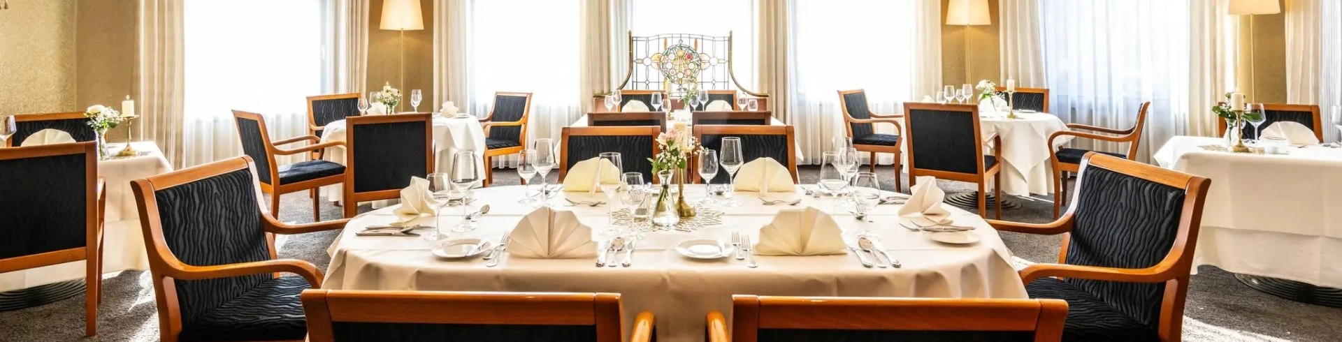 Rund gedeckter Tisch mit weißen Servietten, Gläsern und Besteck in einem hellen Restaurant mit gepolsterten Holzstühlen und großen Fenstern im Schloß-Hotel Petry.