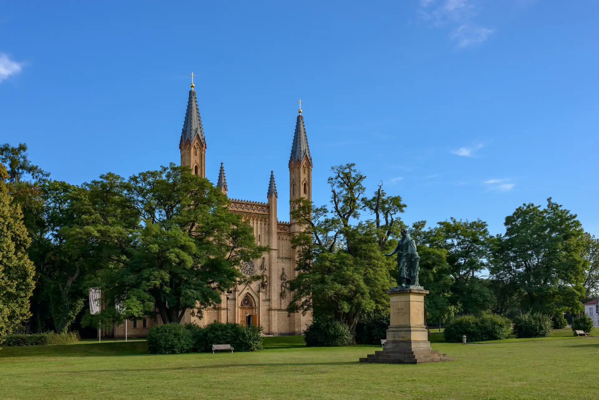 Gotische Kirche mit zwei hohen Türmen im Schlosspark Neustrelitz, umgeben von Bäumen und einer Statue auf einem Sockel im Vordergrund