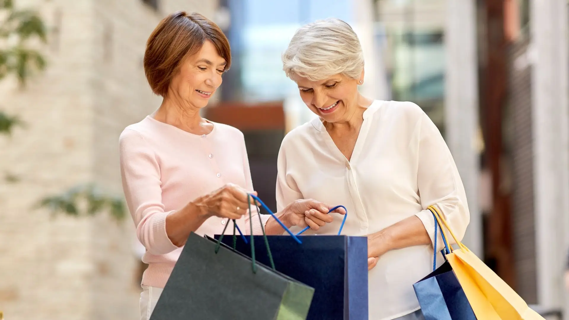 Zwei Frauen mit kurzen Haaren tragen Einkaufstüten in verschiedenen Farben und gehen draußen.