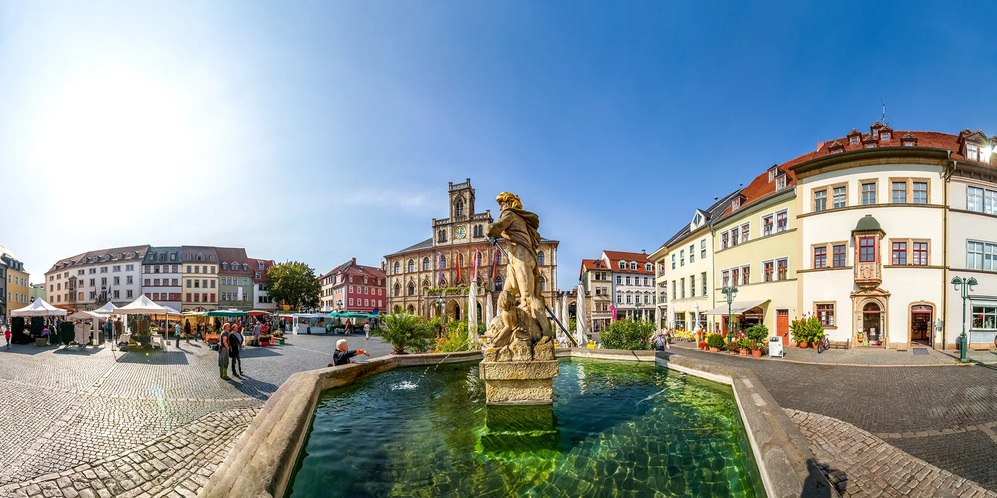 Springbrunnen mit Rathaus und Marktplatz im Hintergrund bei Sonnenschein in der Stadt Weimar